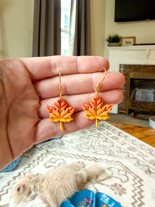 A hand holds a pair of dangling leaf-shaped earrings in front of a living room background with a cat on the floor below.