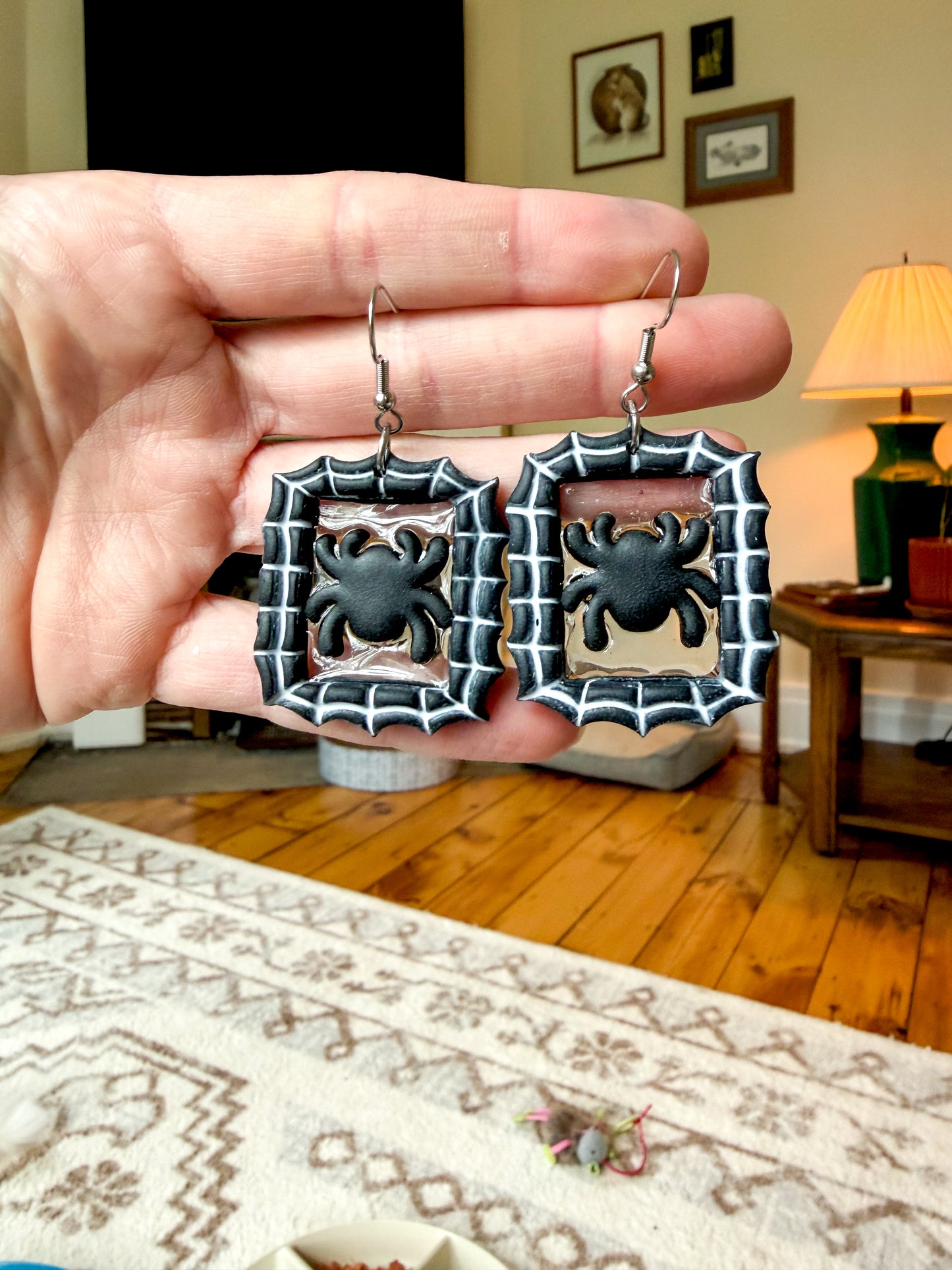 A hand holds a pair of rectangular silver-toned earrings with black spider motifs in the center, shown against a blurred living room background with wood floors and a lamp.