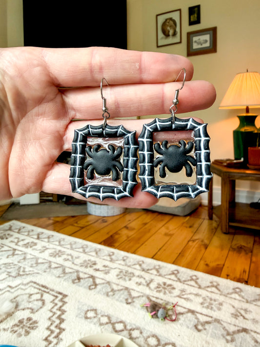 A hand holds a pair of rectangular silver-toned earrings with black spider motifs in the center, shown against a blurred living room background with wood floors and a lamp.