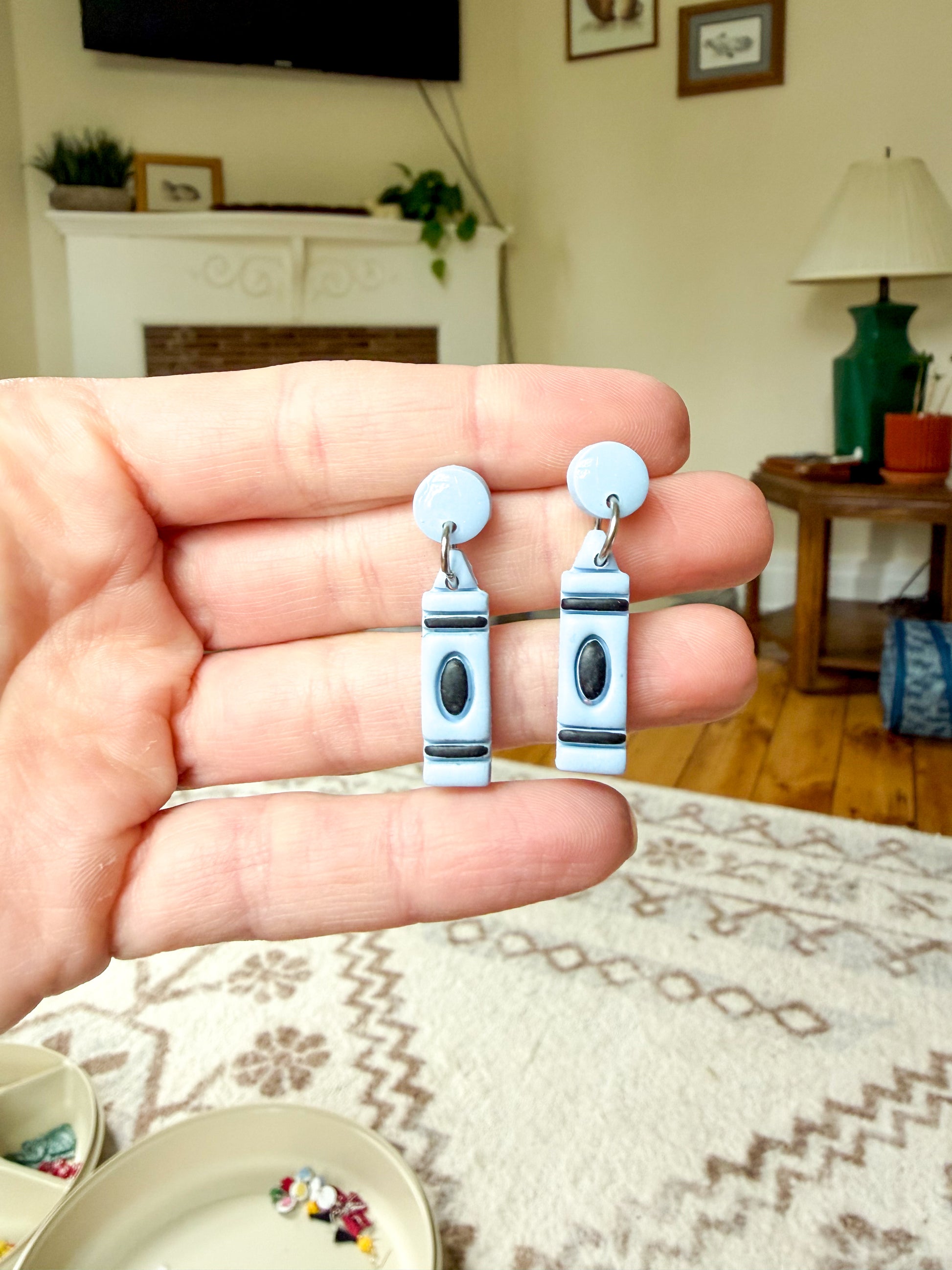 A hand holds a pair of blue polymer clay earrings shaped like crayons, with black and silver details.