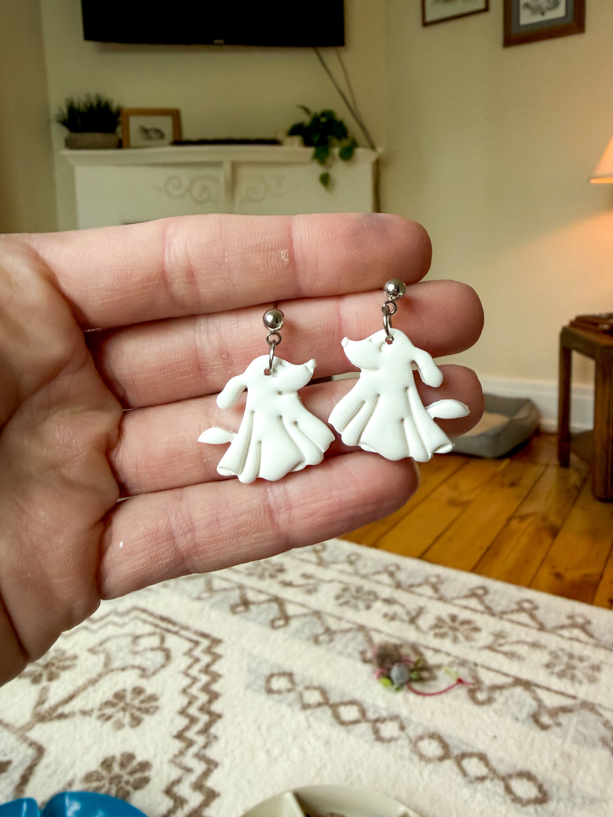 A hand holding a pair of white dog-shaped earrings with silver-toned posts, shown against a blurred living room background.