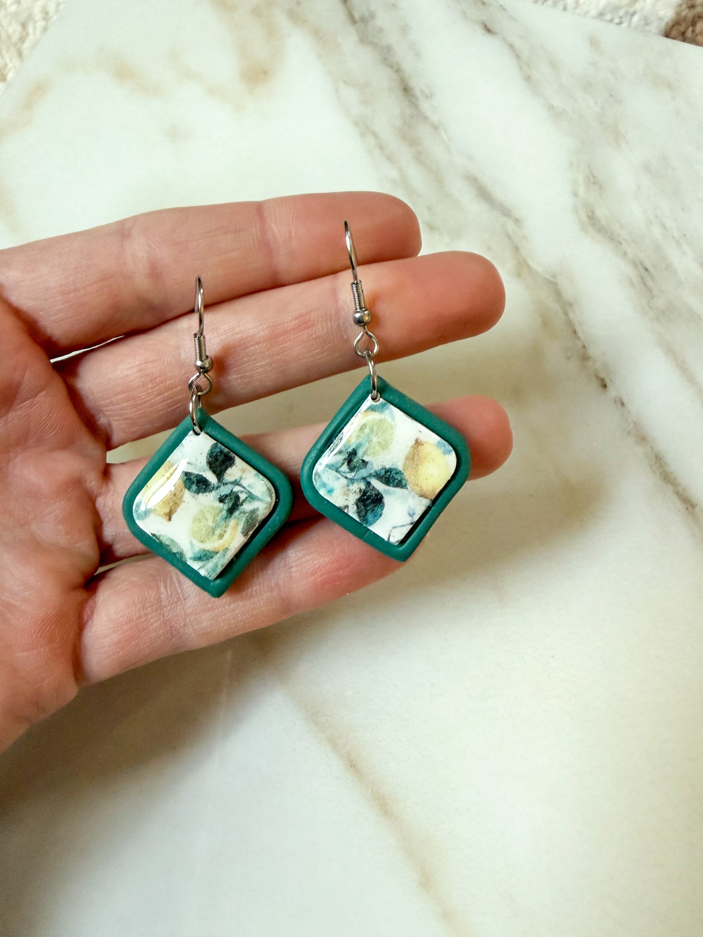 A pair of handmade square drop earrings held in a hand. The earrings feature a dark green border and a central square inlay with a white, green, and yellow lemon pattern, suspended from silver fishhook wires.