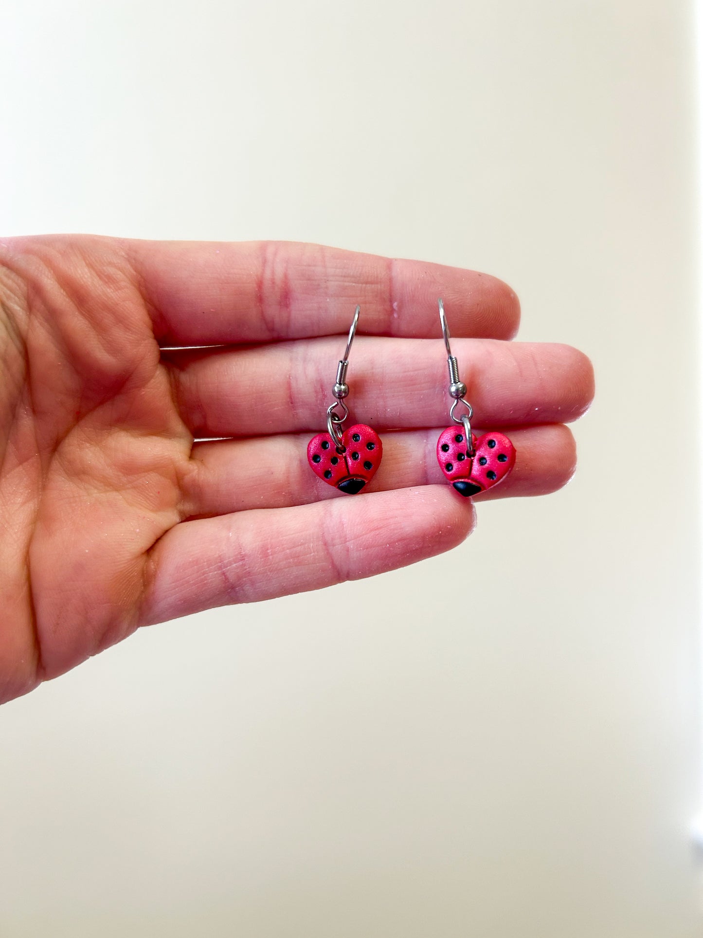 Hand holding a pair of heart shaped earrings that are painted to look like ladybugs ￼