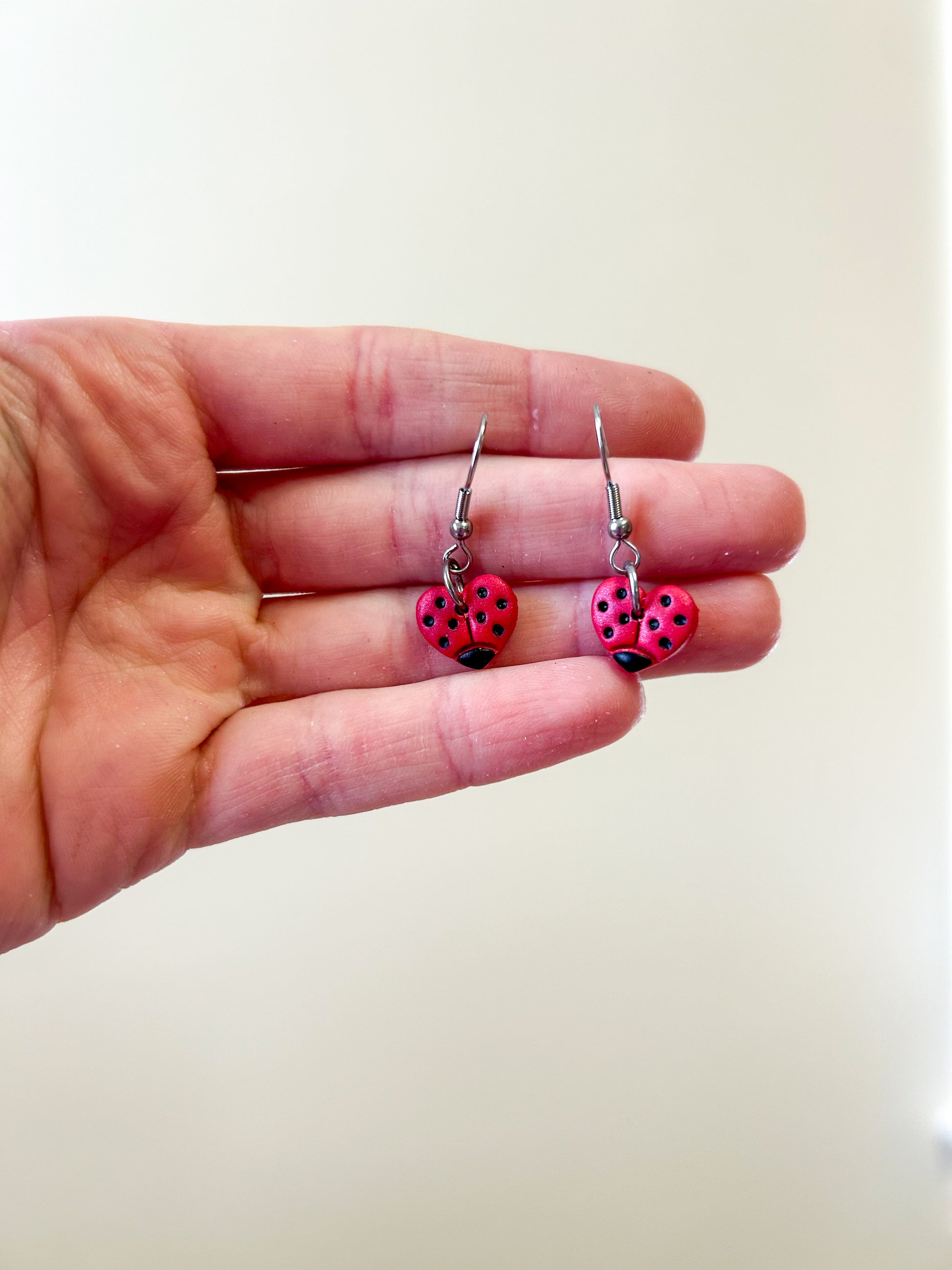 Hand holding a pair of heart shaped earrings that are painted to look like ladybugs ￼