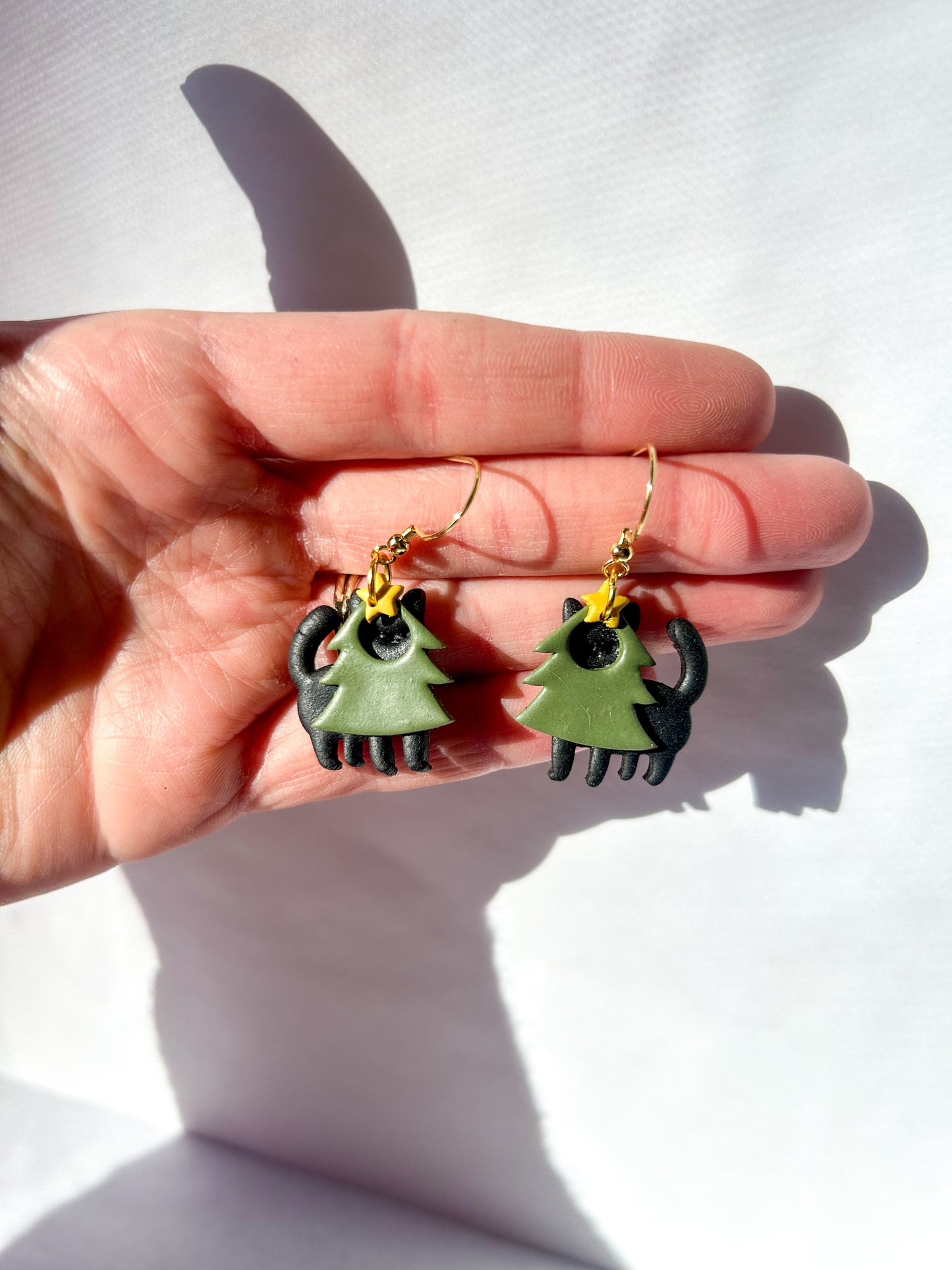 Hand holding a pair of earrings that are black cats in  green tree costumes. Earrings are held against white background.
