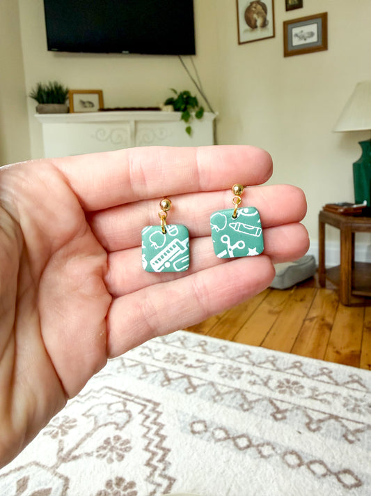 A hand holding a pair of small square earrings with a green and white patterned face and gold-tone posts, shown in a home interior setting.