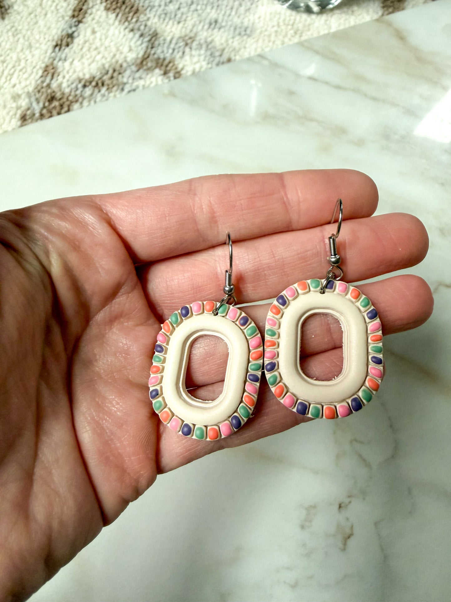 Hand holding two colorful oval earrings with a marble background