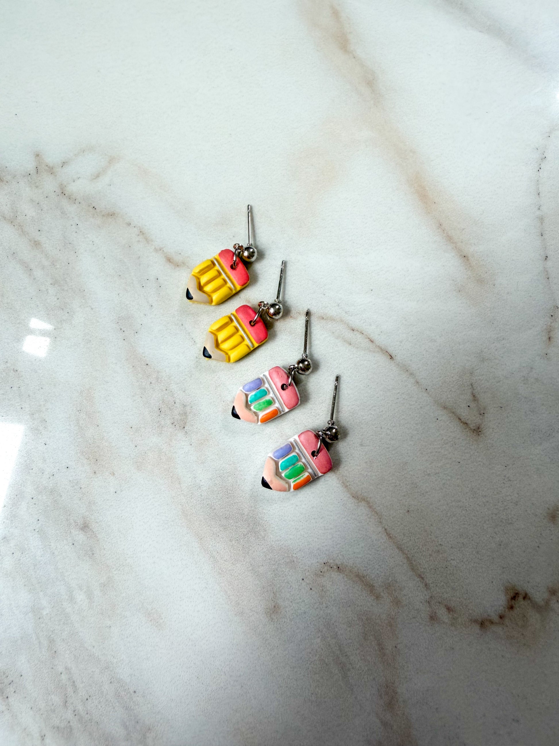 Four colorful pencil-shaped dangle earrings with silver hooks, arranged diagonally on a white marble surface.