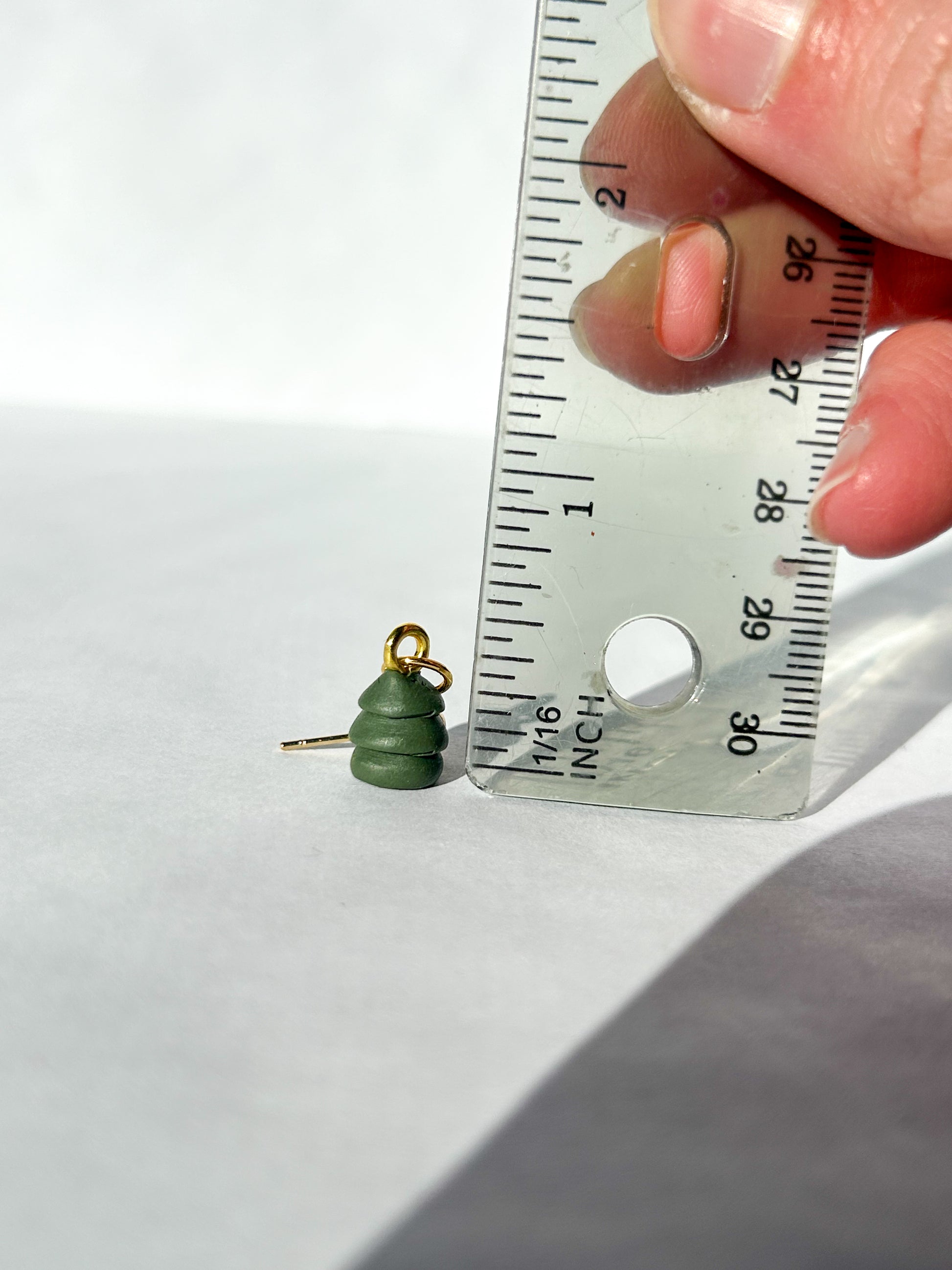 Green Christmas tree-shaped earring being measured with a ruler on a white background. About half an inch in height