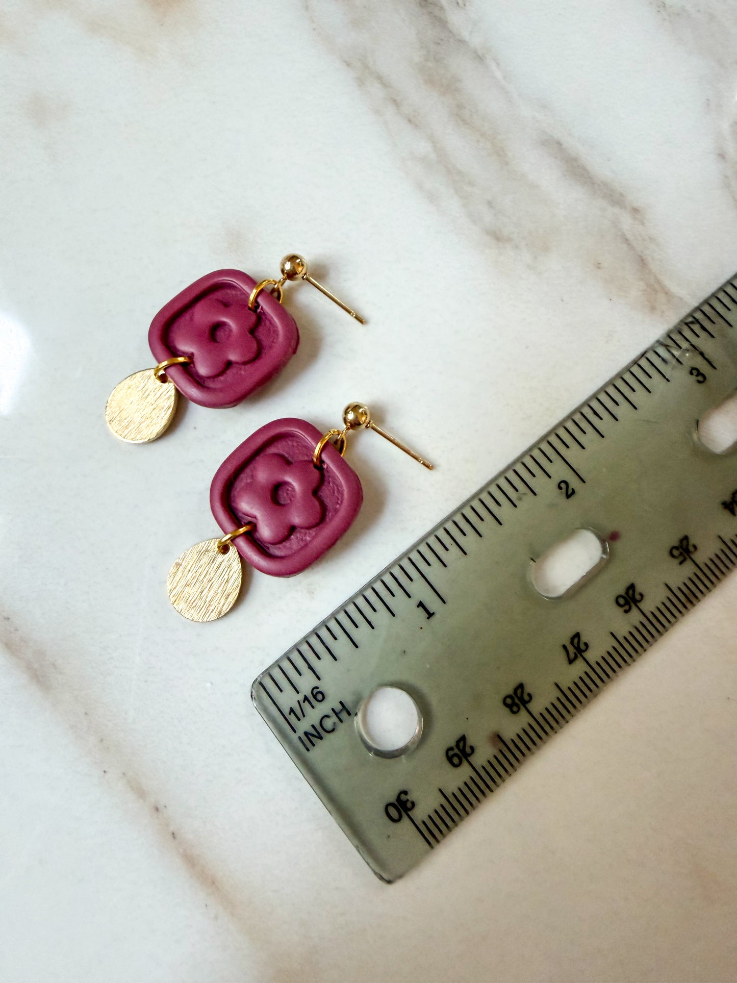 A pair of handmade dangle earrings with magenta pink textured square charms and gold-tone disc charms, displayed next to a ruler on a light marble surface. 1 1/2 inches in length. 