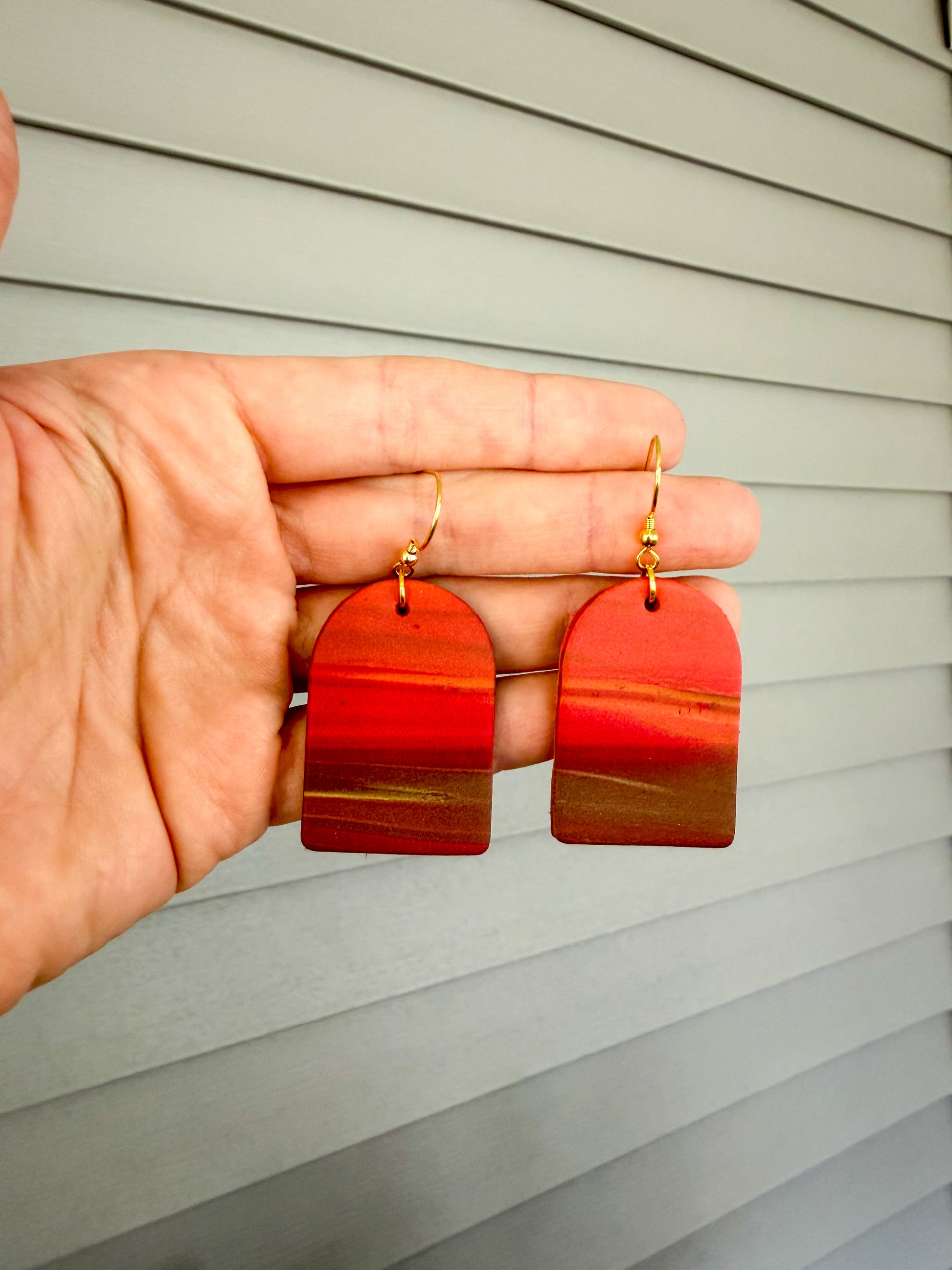 Earrings held in a hand against a house. The design resembles a sunset with colors fading from orange to brown. 