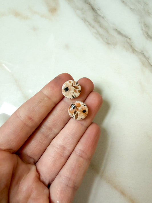 A close-up of a hand holding two small round stud earrings with a floral pattern on a light marble surface.