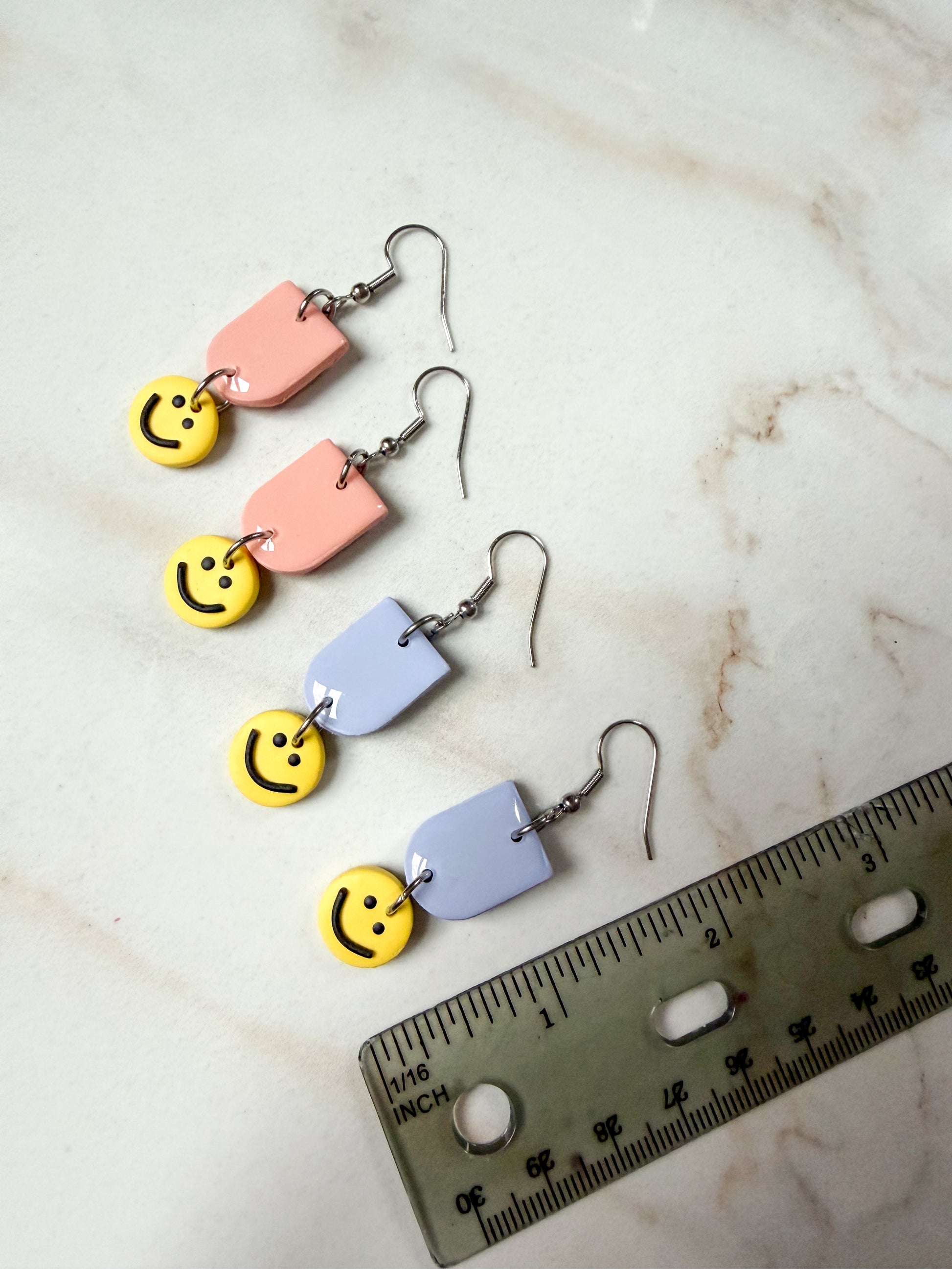 Colorful smiley face earrings on a marble surface with a ruler for scale. About 2 inches long.