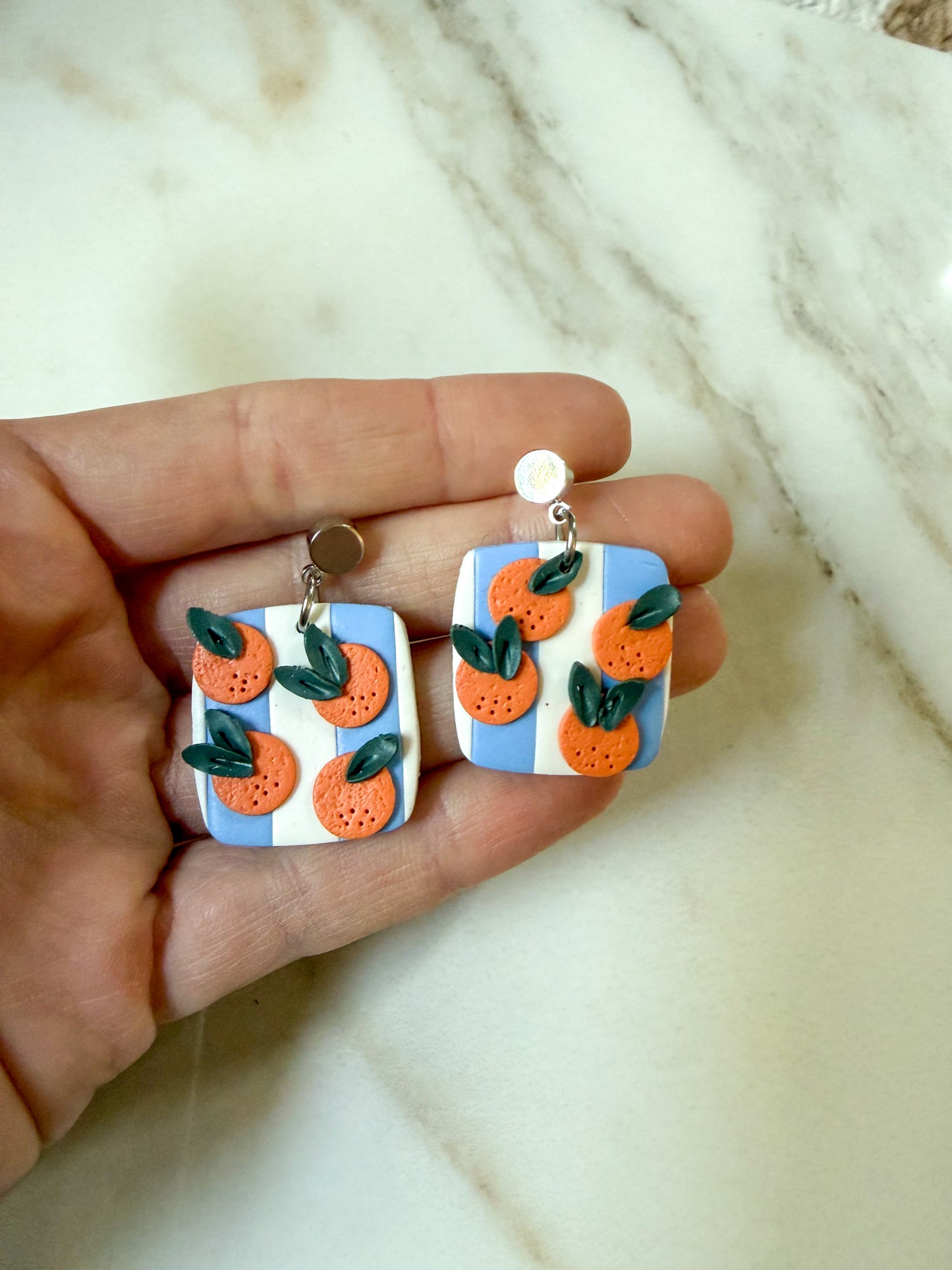 A pair of handmade rectangular drop earrings with a blue and white striped background, decorated with orange citrus slices and green leaves, held in a person's hand against a marble surface.