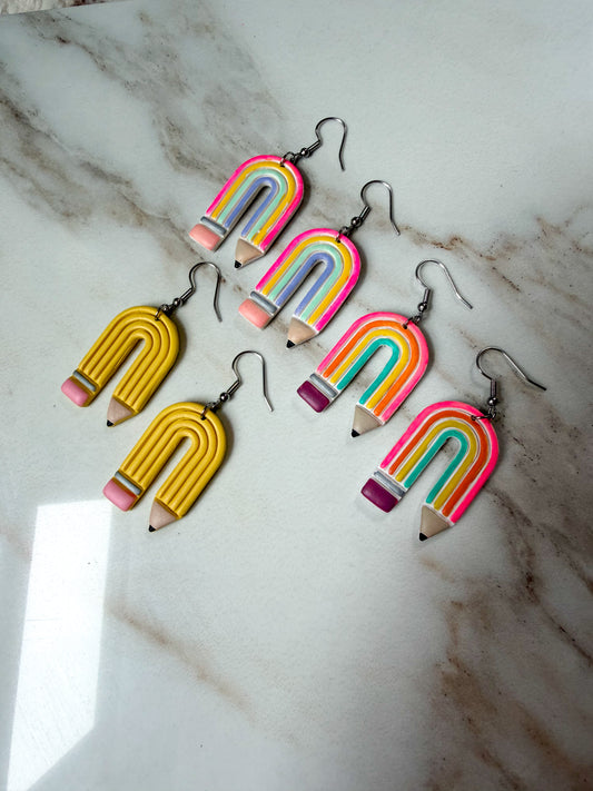 Six rainbow pencil earrings with silver-tone fishhook wires are arranged on a white and gray marble surface. Two earrings are yellow with pink erasers, and four have rainbow stripes in blue, green, orange, pink, and yellow.