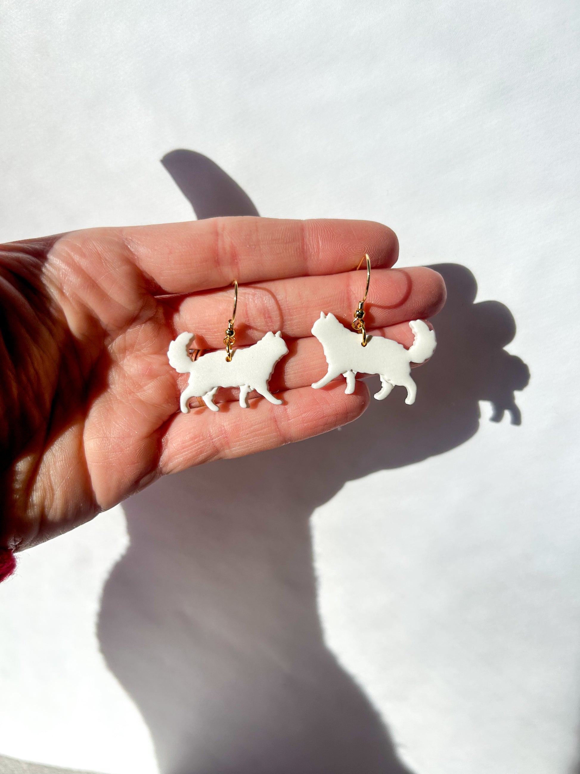 Hand holding two white long haired cat-shaped earrings against a light background