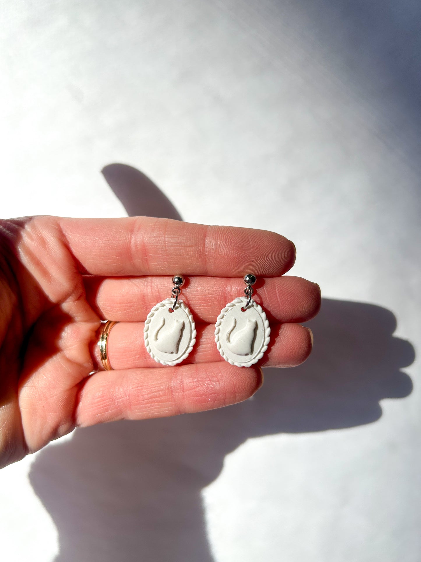 White earrings held in a hand against a white background. The earrings resemble a scalloped frame with a raised cat silhouette in the center.