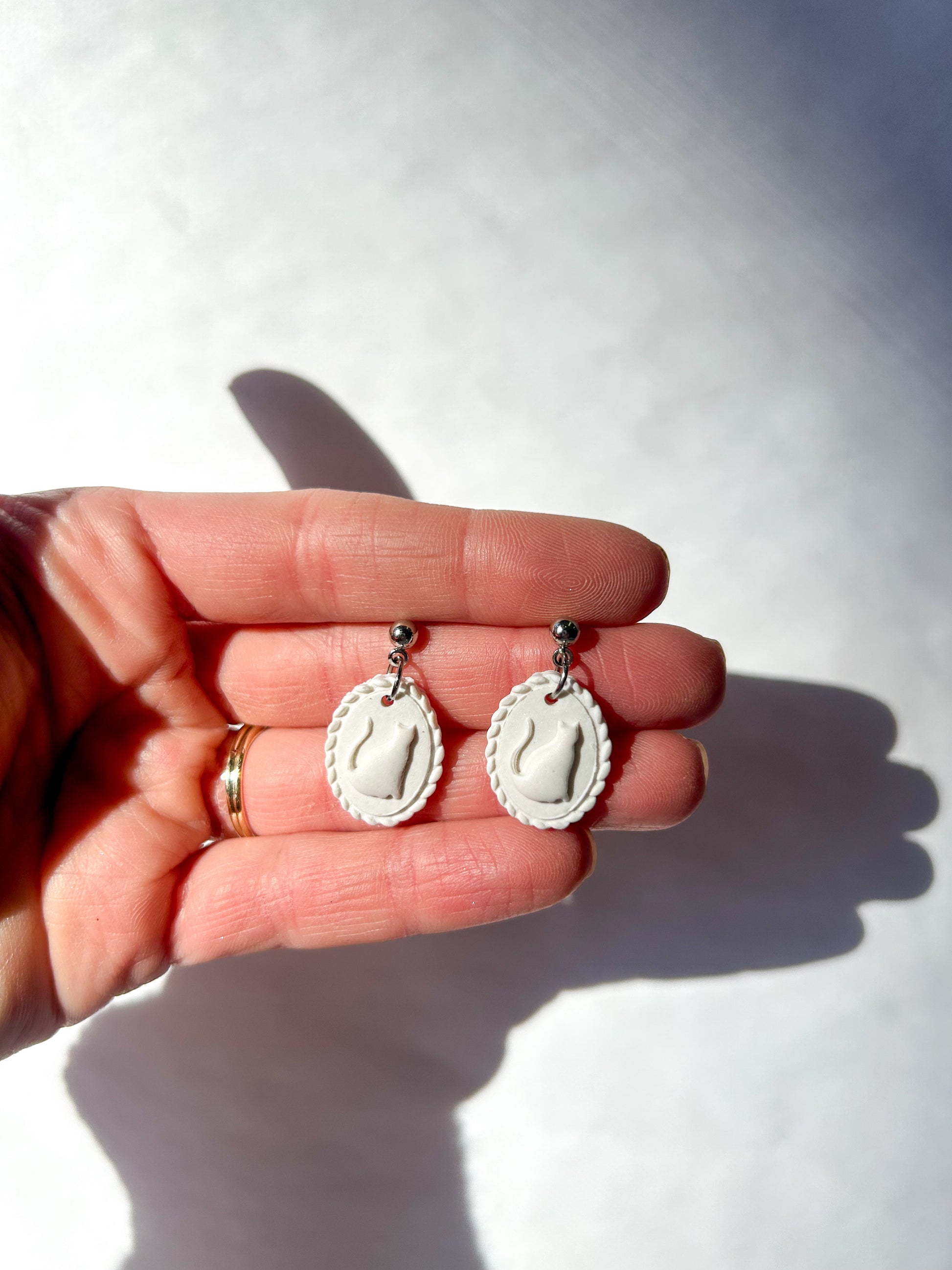 White earrings held in a hand against a white background. The earrings resemble a scalloped frame with a raised cat silhouette in the center.