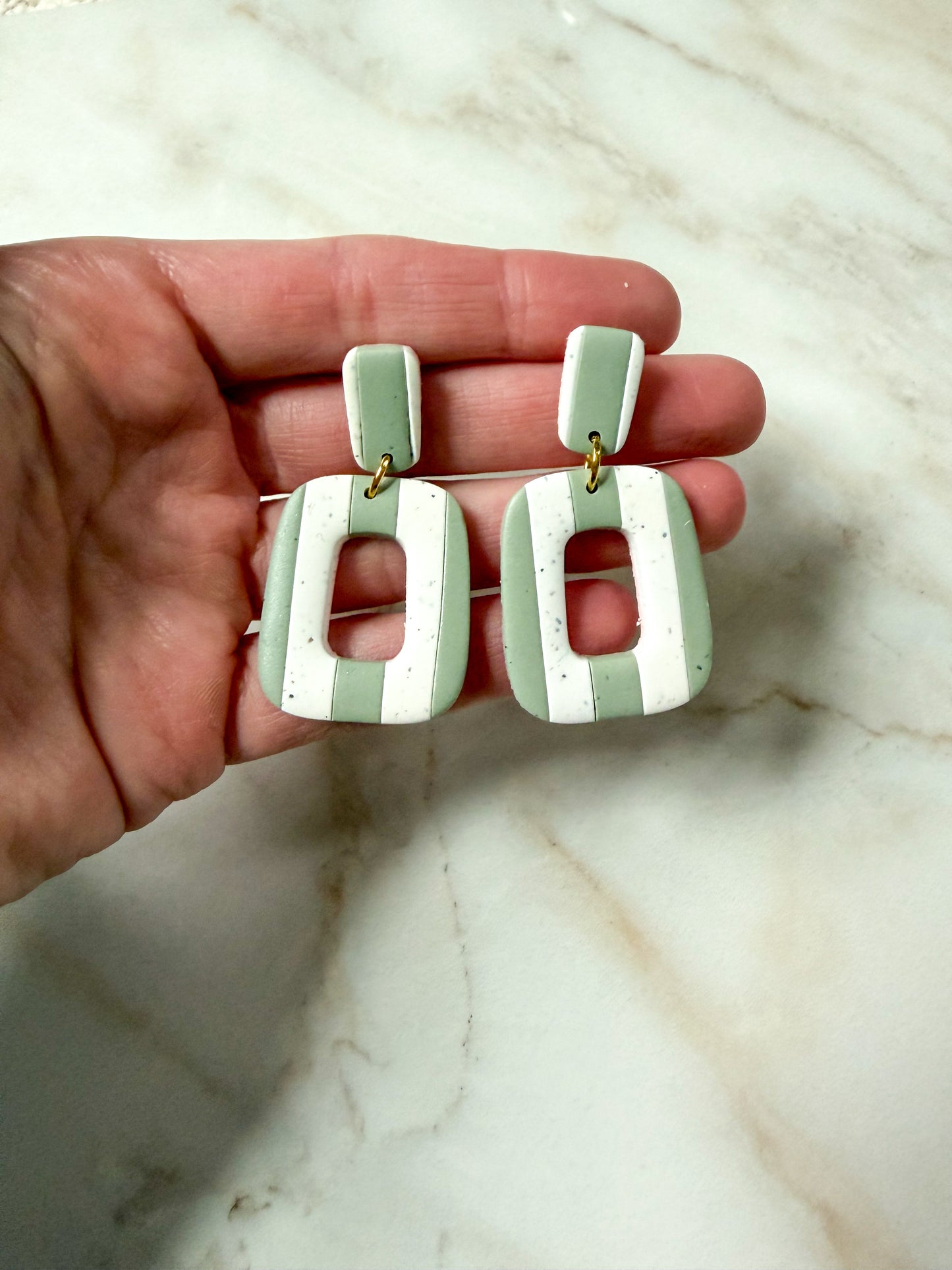 Hand holding a pair of green and white striped earrings on a marble surface