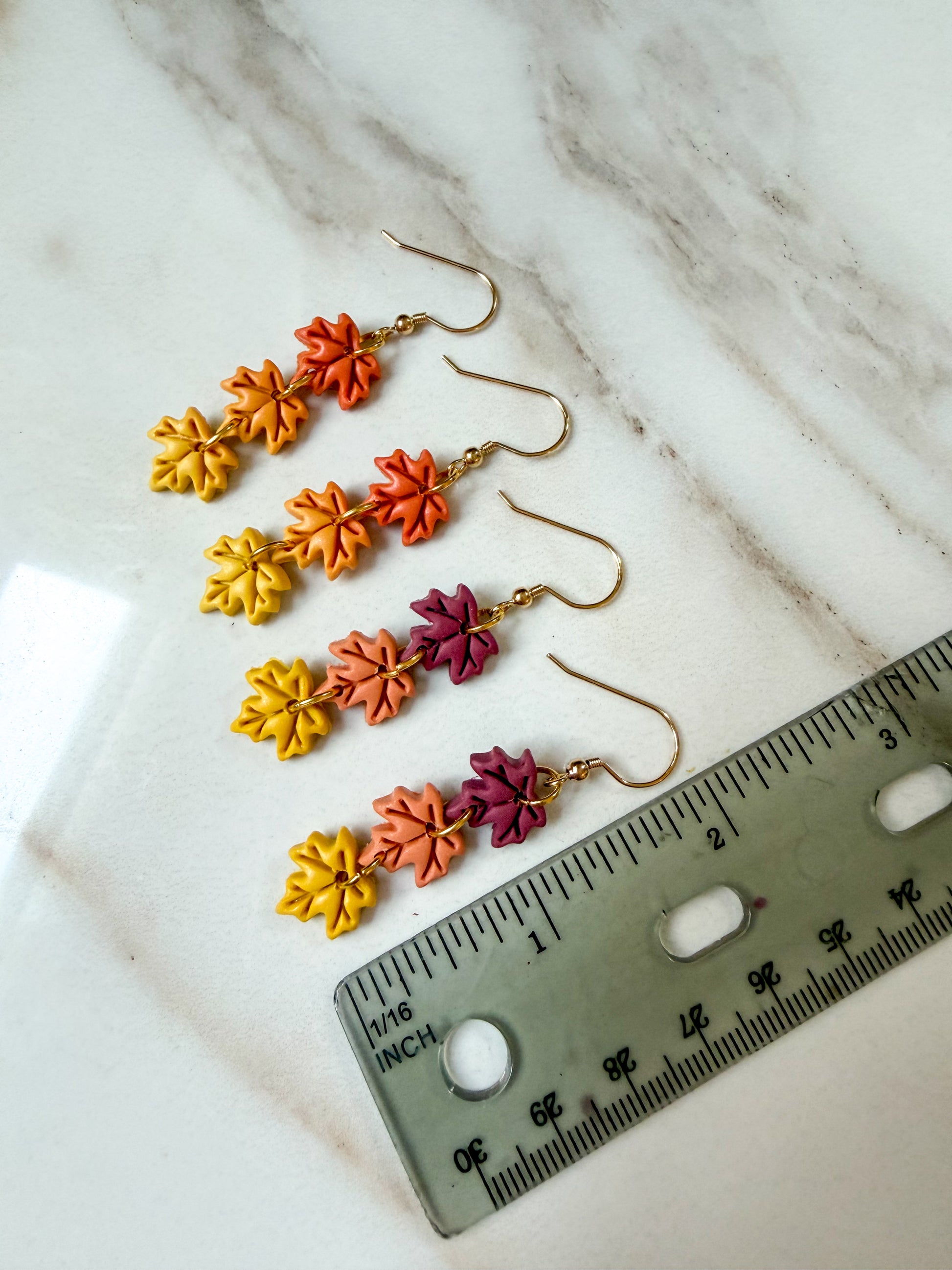 Two pairs of dangling earrings with small leaf-shaped charms in yellow, orange, and burgundy, displayed on a white marble surface next to a ruler for scale. About 2 inches long.