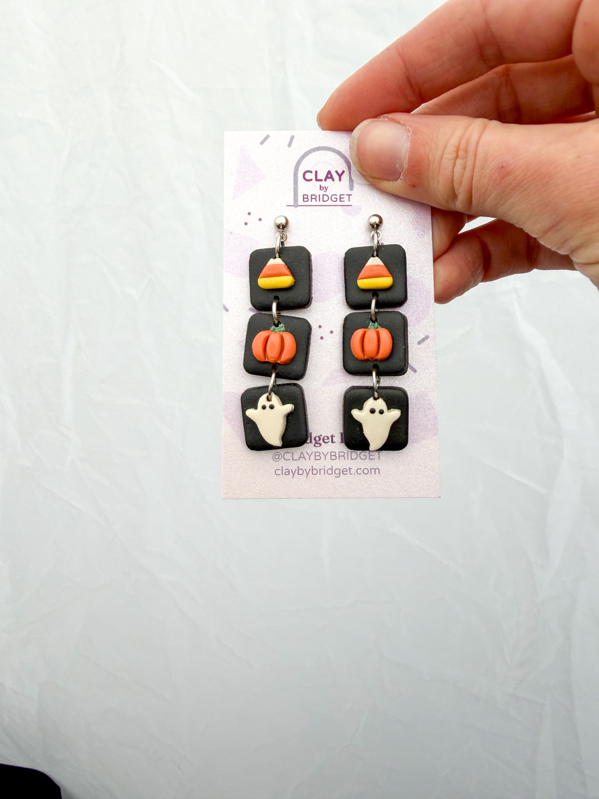A hand holds a small display card featuring a pair of Halloween-themed dangle earrings. The earrings have black square charms with candy corn, orange pumpkins, and white ghosts, suspended from silver hooks.