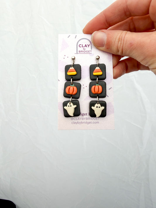 A hand holds a small display card featuring a pair of Halloween-themed dangle earrings. The earrings have black square charms with candy corn, orange pumpkins, and white ghosts, suspended from silver hooks.