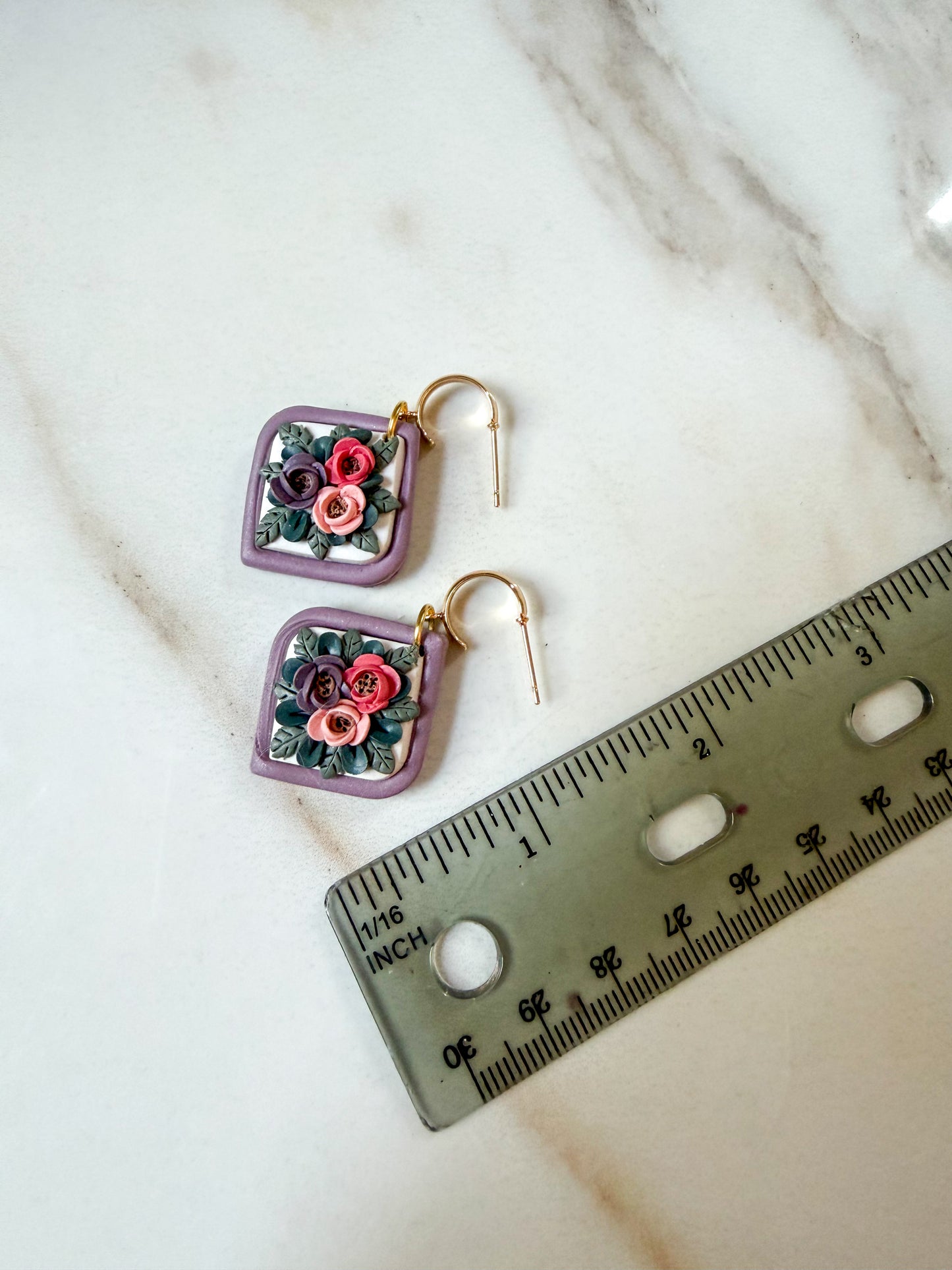A pair of square dangle earrings with purple borders and colorful floral details, displayed on a white marble surface next to a ruler for scale. An inch and a half in length. 