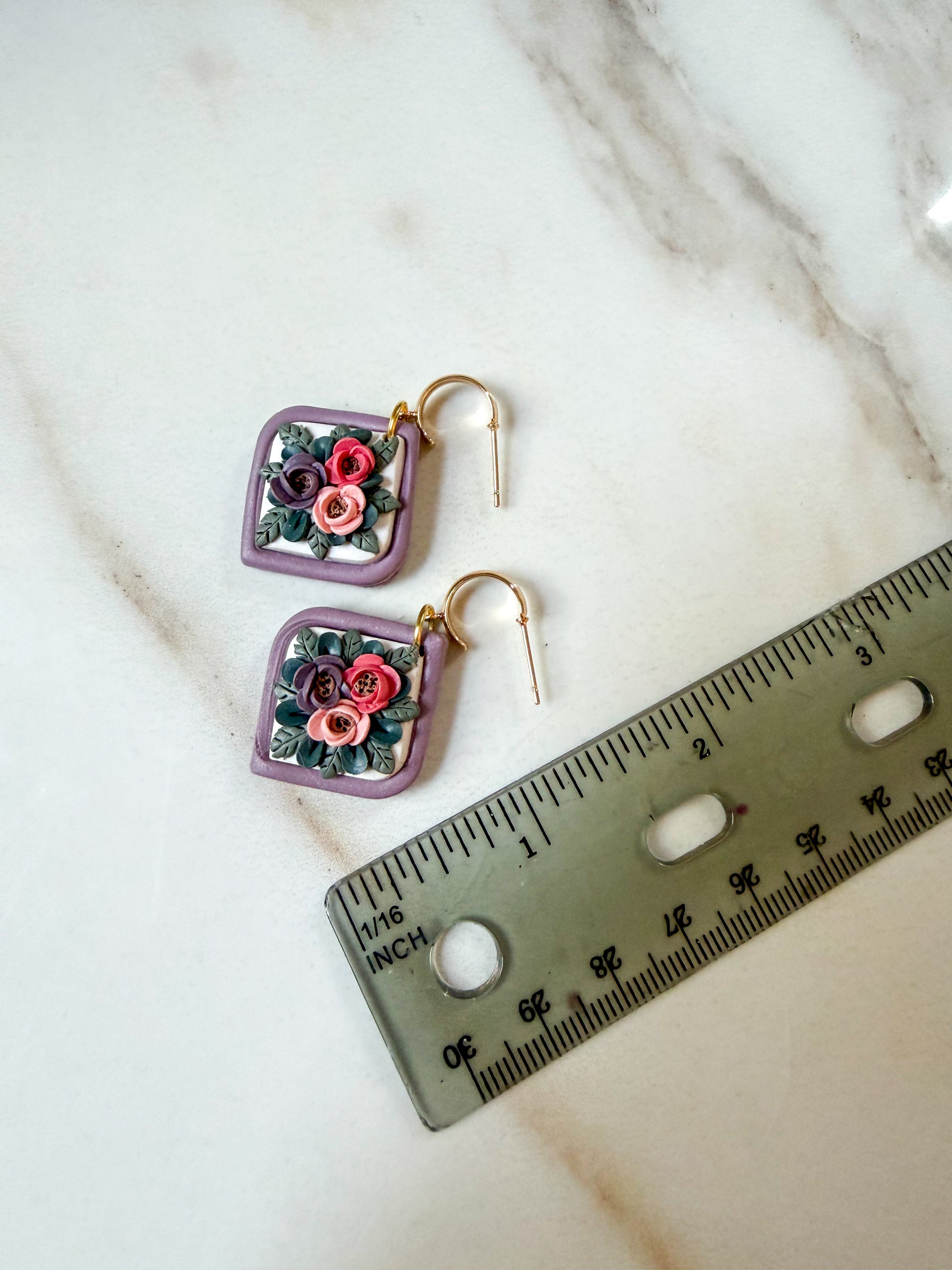 A pair of square dangle earrings with purple borders and colorful floral details, displayed on a white marble surface next to a ruler for scale. An inch and a half in length. 