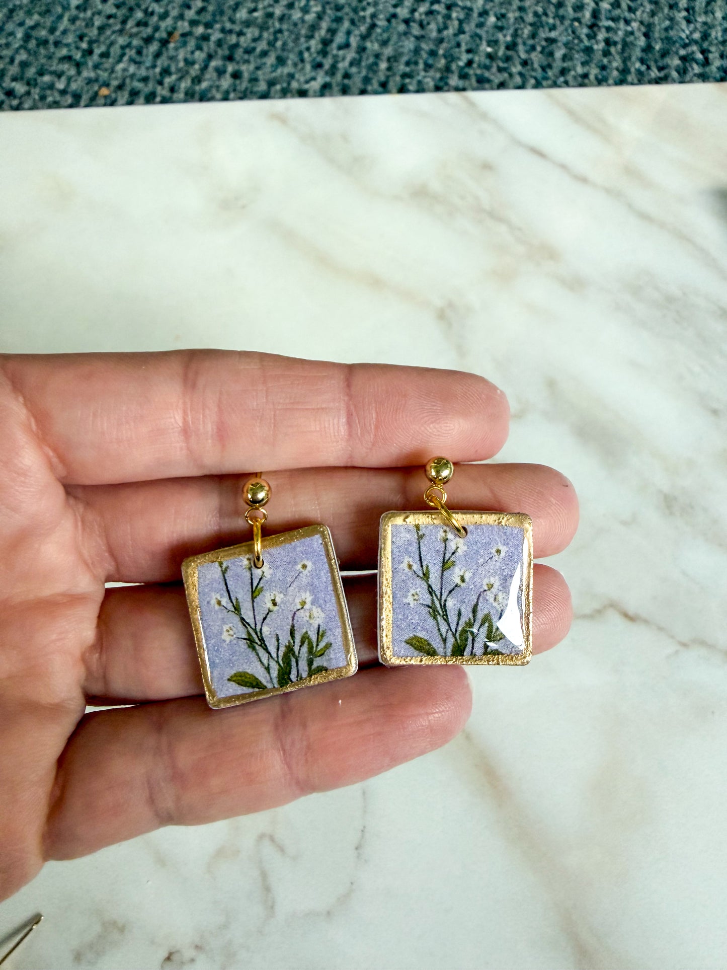Square earrings with floral design held between fingers on a marble surface