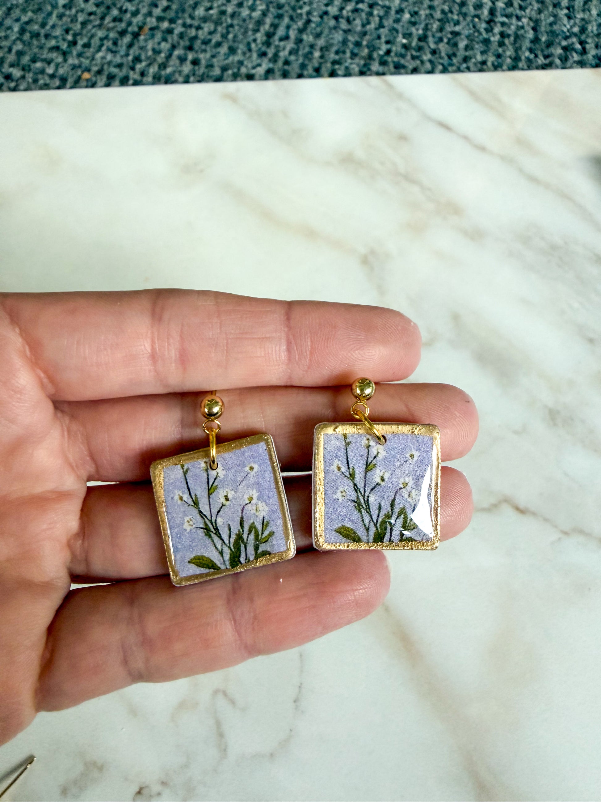 Square earrings with floral design held between fingers on a marble surface