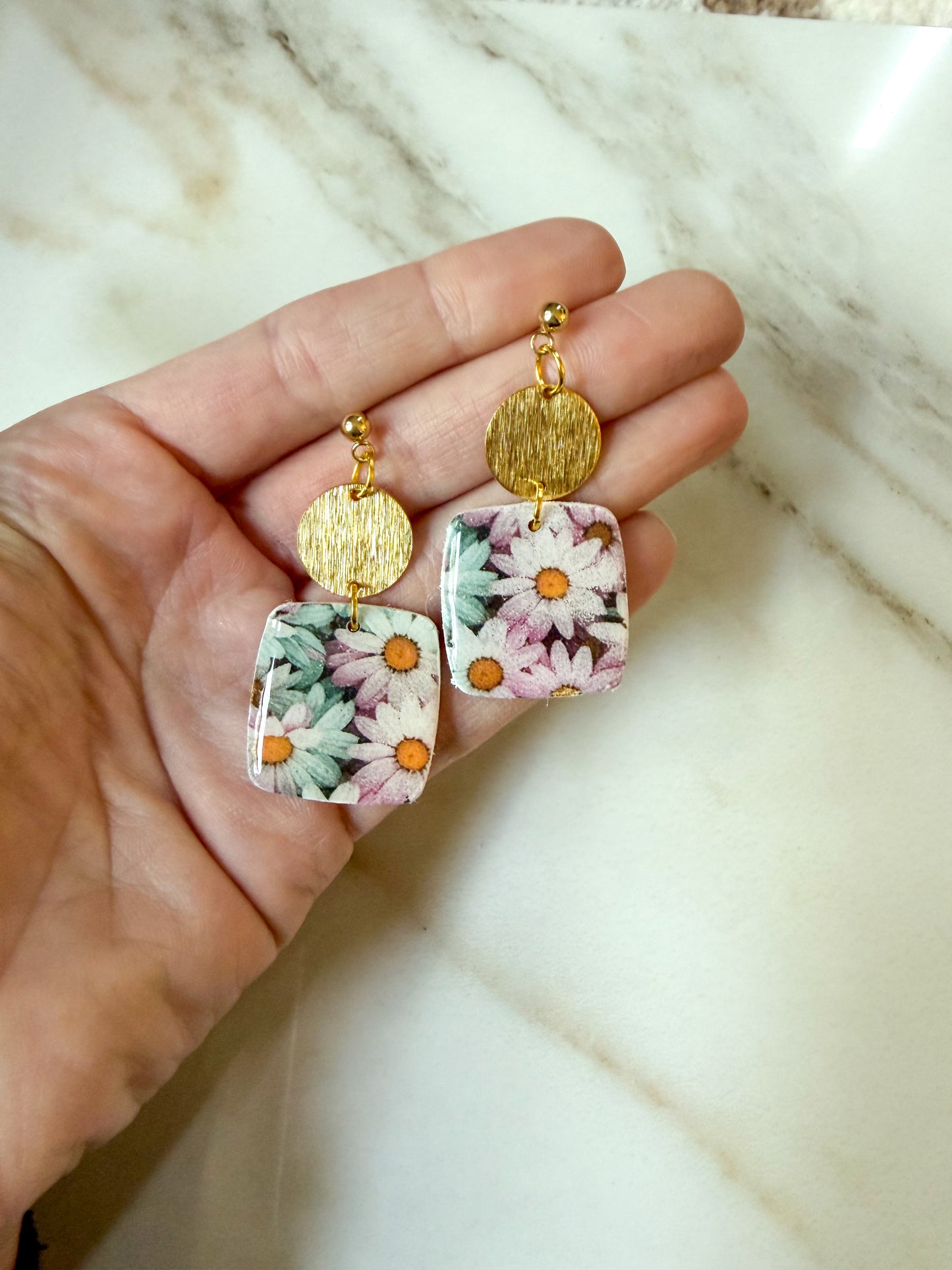 A pair of dangling earrings held in a hand, featuring square floral resin pendants with pink, white, and orange flowers, gold-tone round discs, and gold-tone hooks, set against a light marble background.