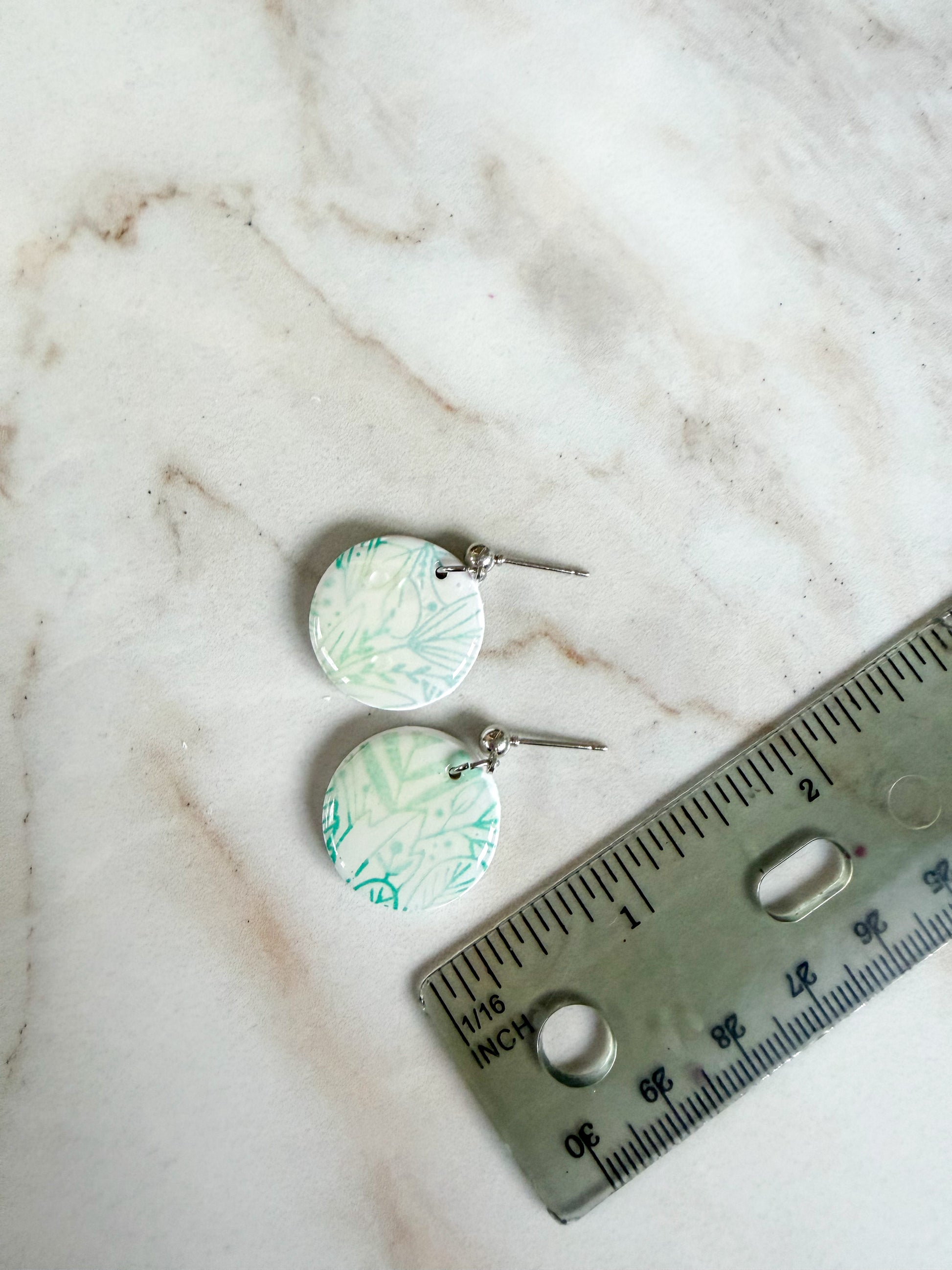 Pair of earrings with a green and white pattern on a marble surface with a ruler for scale. About one inch in length 