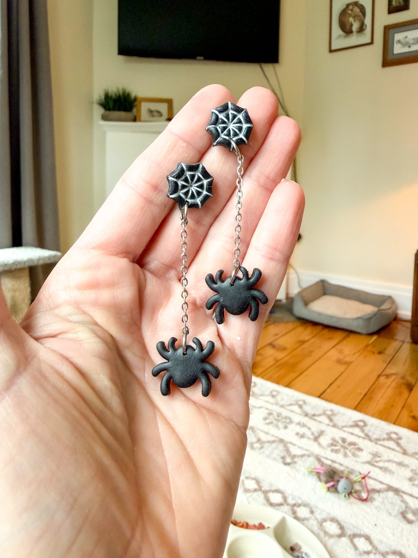 A hand holds a pair of dangling earrings in a home interior. The earrings have spiderweb-shaped studs connected by short chains to black spider charms.