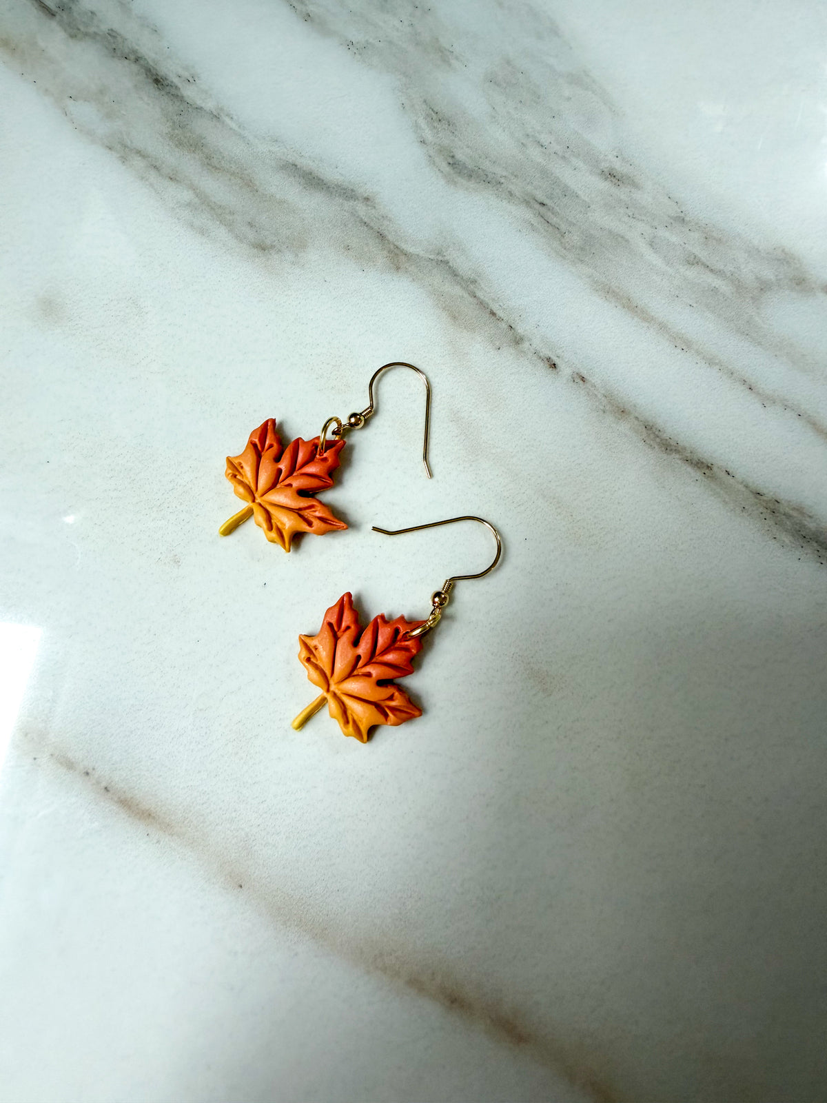 A pair of dangling earrings shaped like orange and yellow autumn maple leaves, suspended from gold fishhook wires, displayed on a white marble surface.
