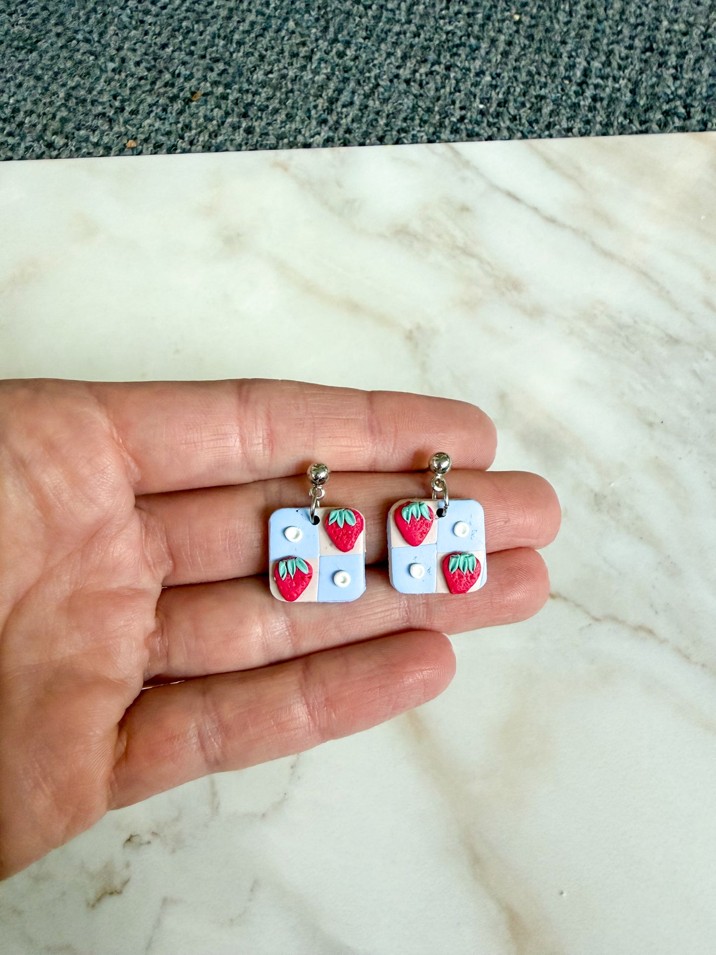 Hand holding two square-shaped earrings with strawberry designs on a marble surface.