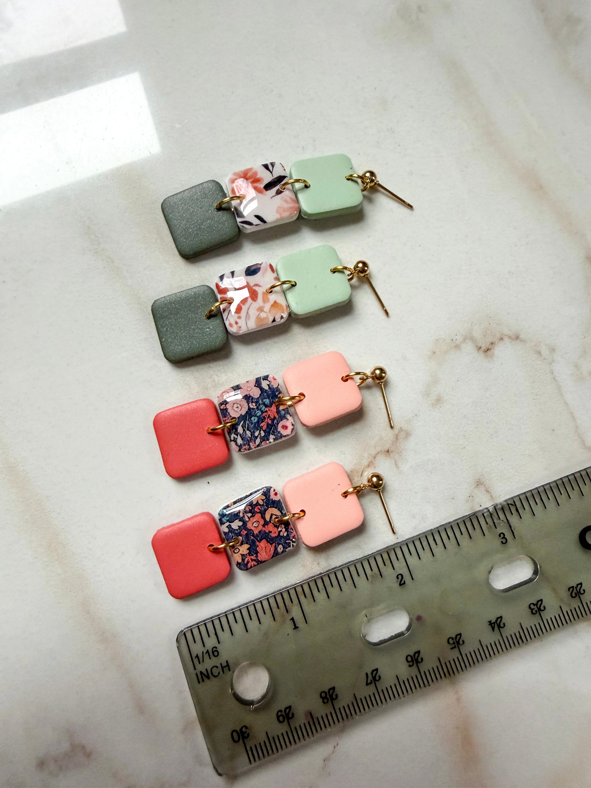 Two pairs of earrings with floral and solid square designs on a marble surface with a ruler for scale. About 2 inches long. 