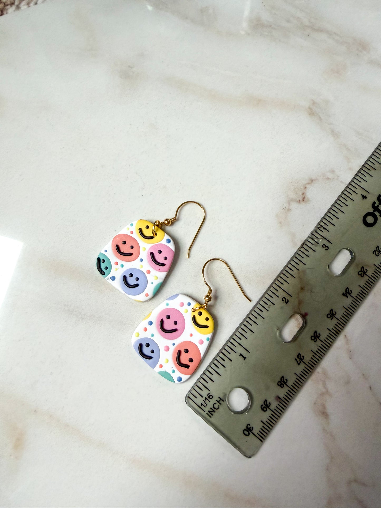 Colorful smiley face earrings on a marble surface with a ruler for scale. About 2 inches long