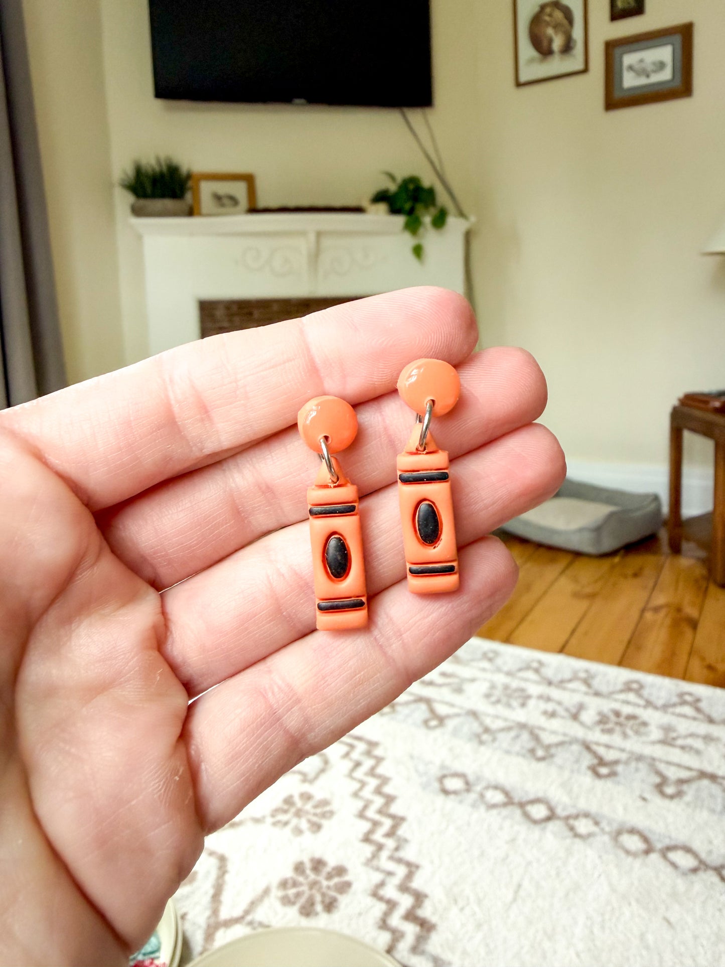 A hand holds a pair of orange polymer clay earrings shaped like crayons, with black and silver details.