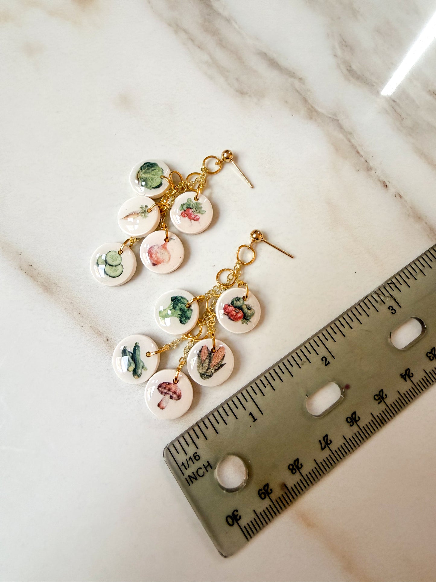 A pair of dangling earrings with small round white charms printed with colorful vegetable illustrations, attached to gold-tone hooks. A ruler is placed beside them on a light marble surface for scale. Almost 3 inches in length. 