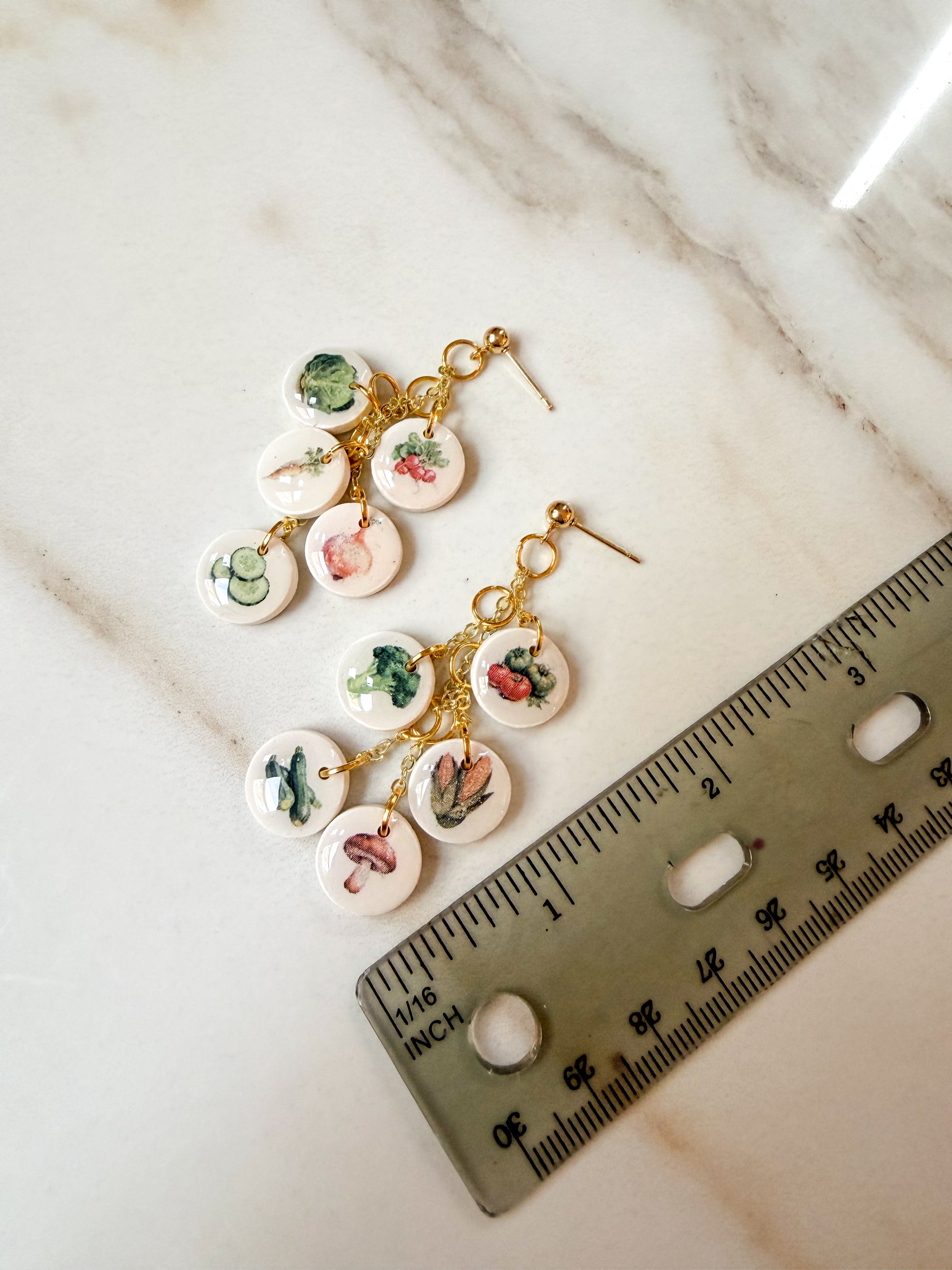 A pair of dangling earrings with small round white charms printed with colorful vegetable illustrations, attached to gold-tone hooks. A ruler is placed beside them on a light marble surface for scale. Almost 3 inches in length. 