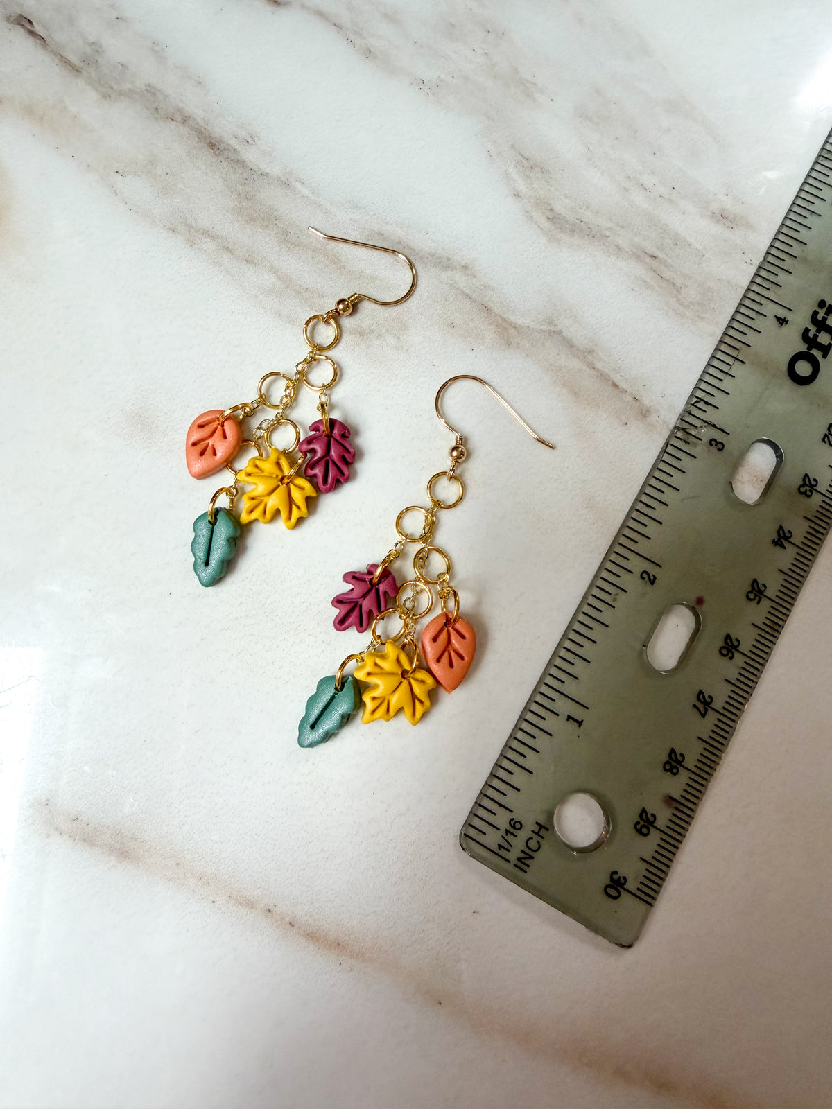 A pair of gold-tone dangle earrings with colorful autumn leaf charms in orange, yellow, pink, and teal, displayed on a white marble surface next to a ruler for scale. Just under 3 inches in length. 