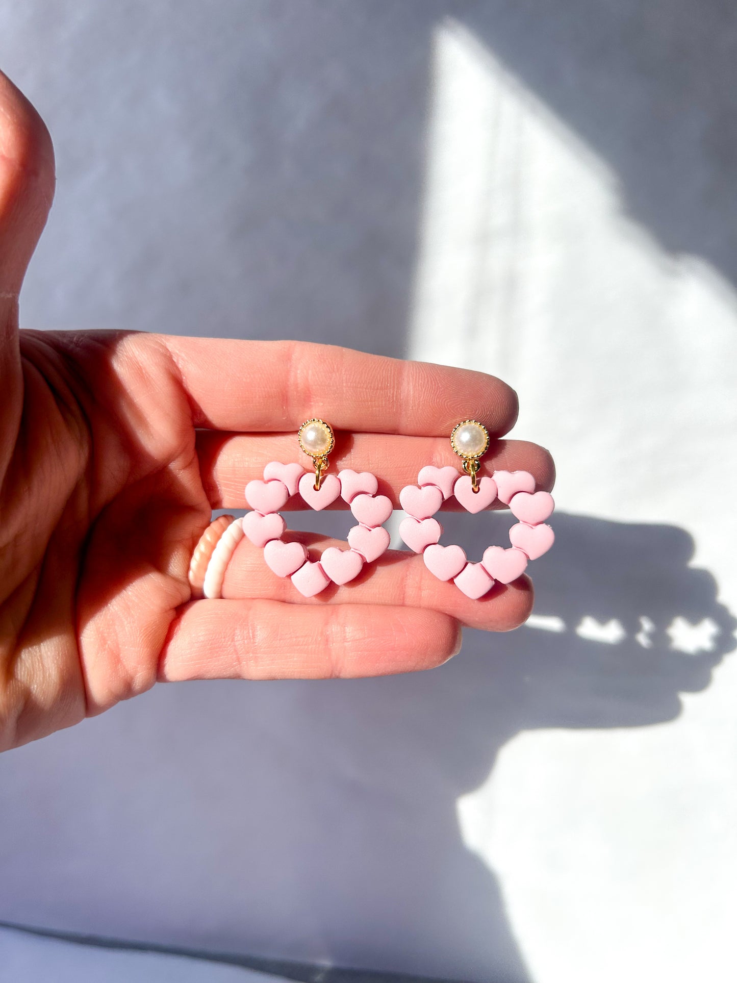 Heart-shaped earrings held in a hand with a neutral background. Posts are pearls