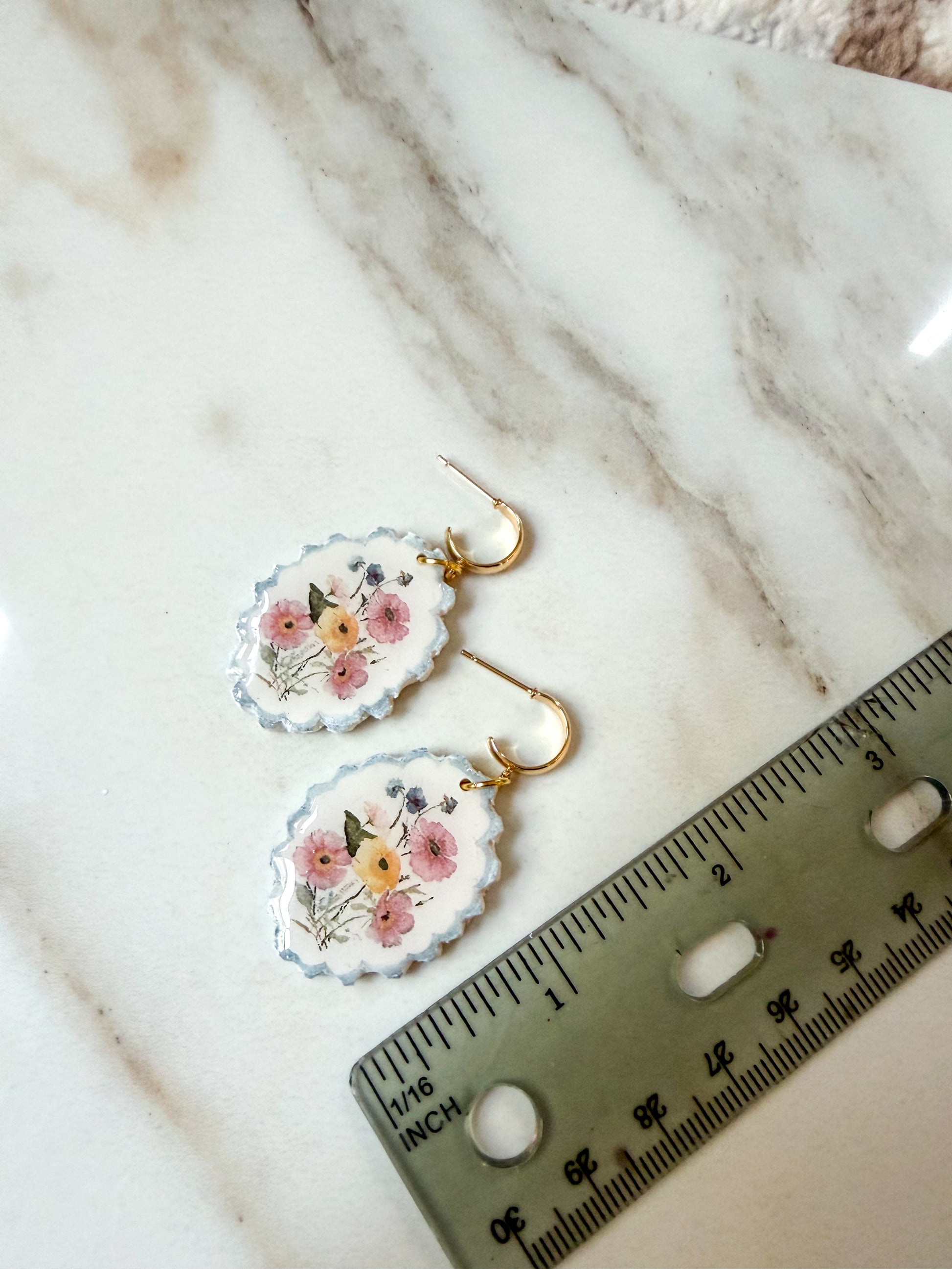 A pair of scalloped oval earrings with a delicate pink and yellow floral design on a white background, suspended from gold hooks. They are displayed on a white marble surface next to a metal ruler for scale. About 2 inches long. 