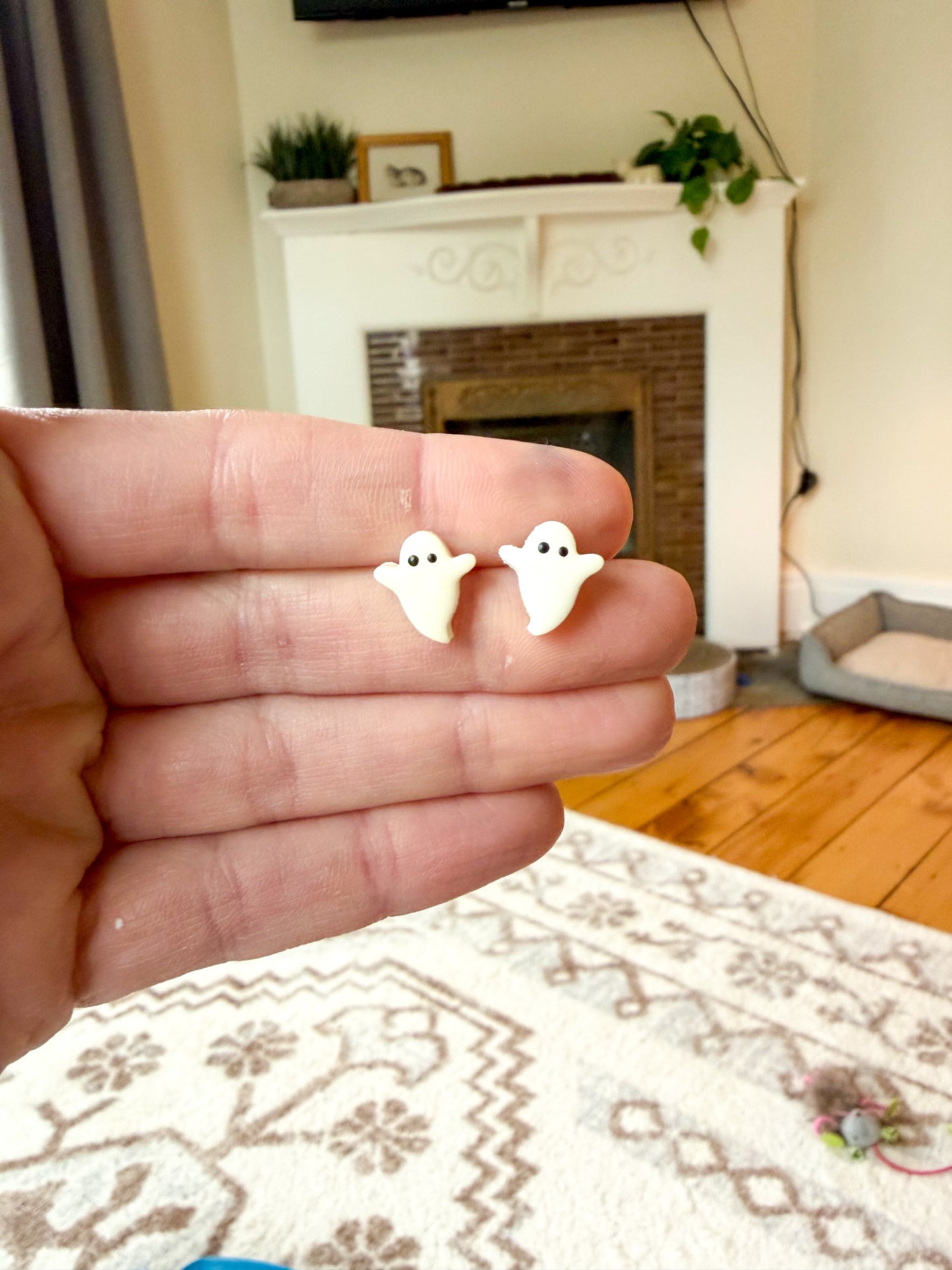 A hand holding a pair of small white ghost-shaped stud earrings in front of a living room background with a fireplace and patterned rug.