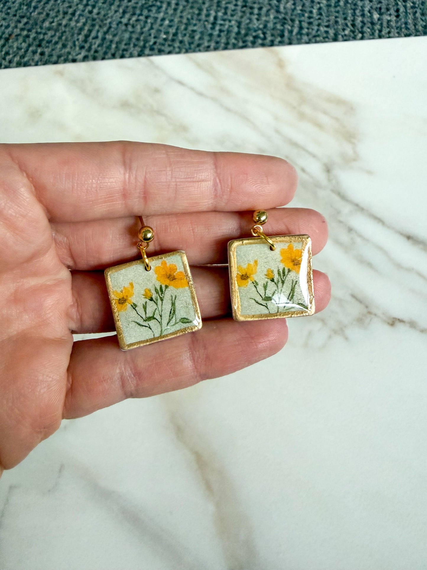 Square earrings with floral design held between fingers on a marble surface