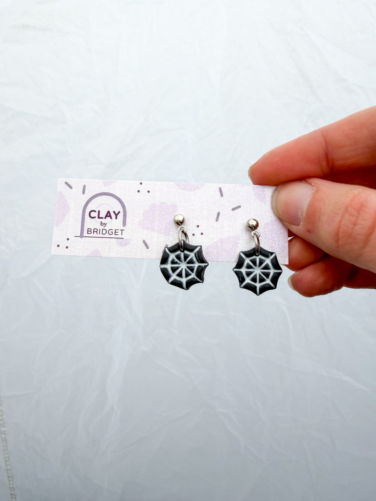 Small black earrings with a white spiderweb design. Held in a hand against a white surface. 