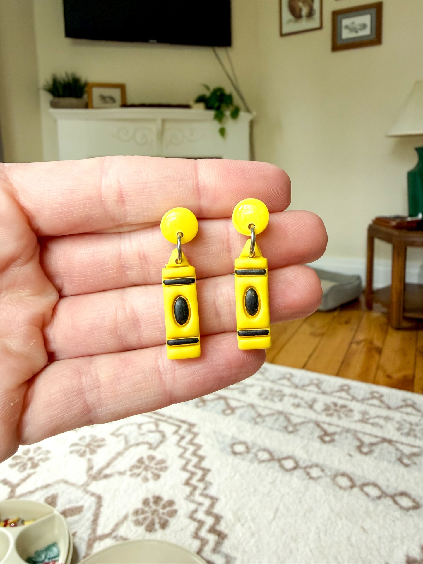 A hand holds a pair of yellow polymer clay earrings shaped like crayons, with black and silver details.