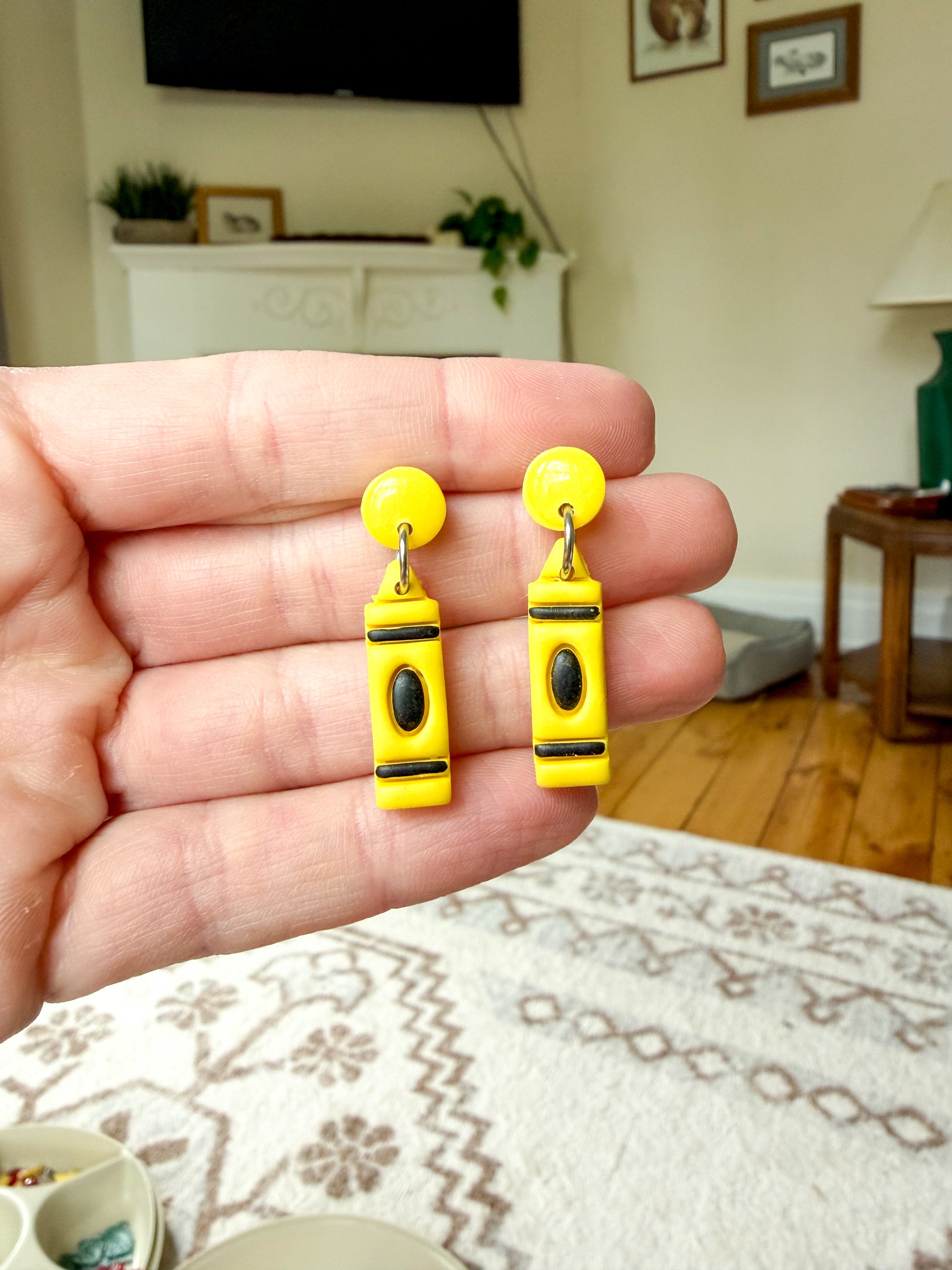 A hand holds a pair of yellow polymer clay earrings shaped like crayons, with black and silver details.