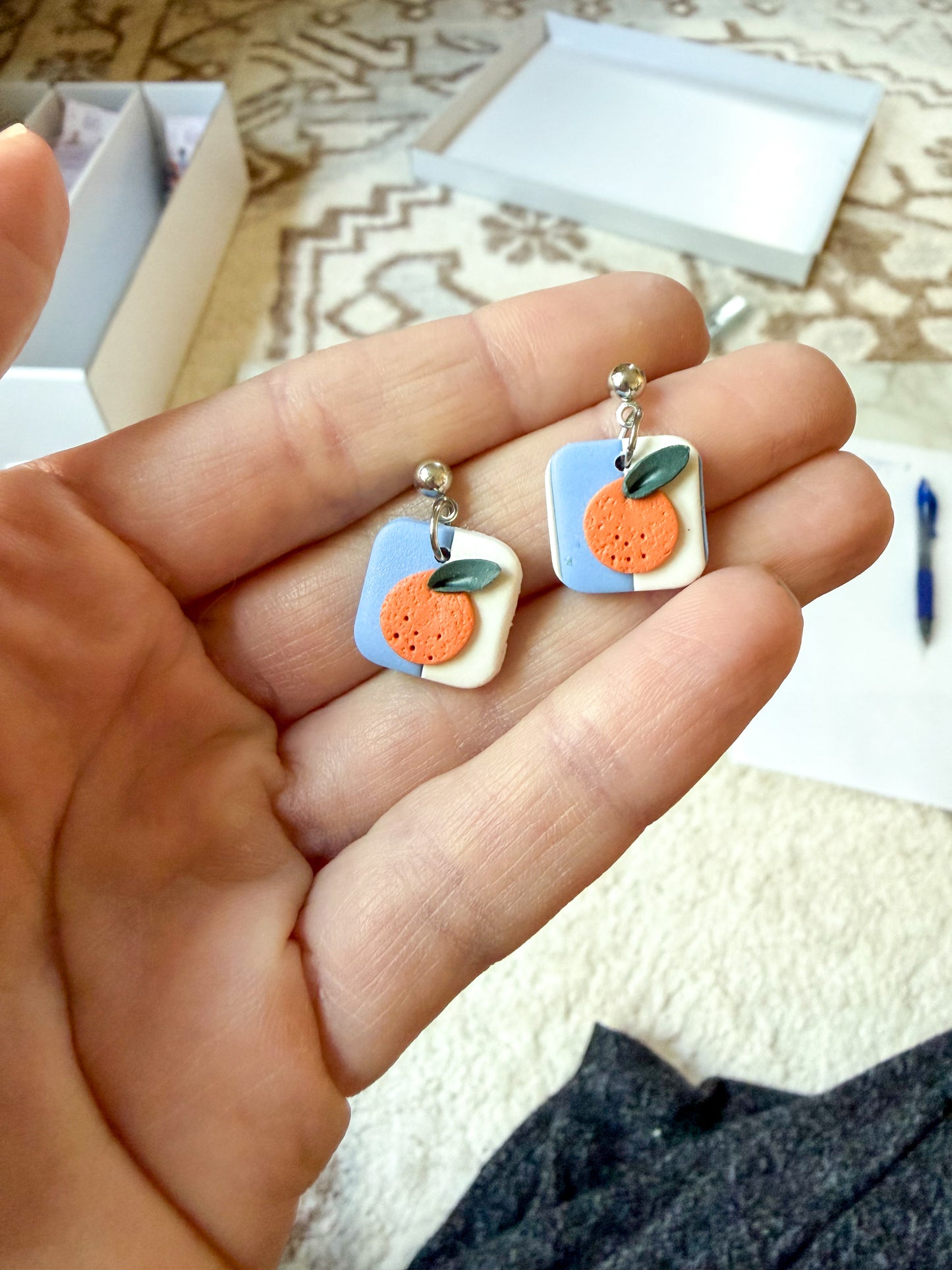 A close-up of a hand holding a pair of small square earrings with orange citrus fruit charms on a light blue and white background.