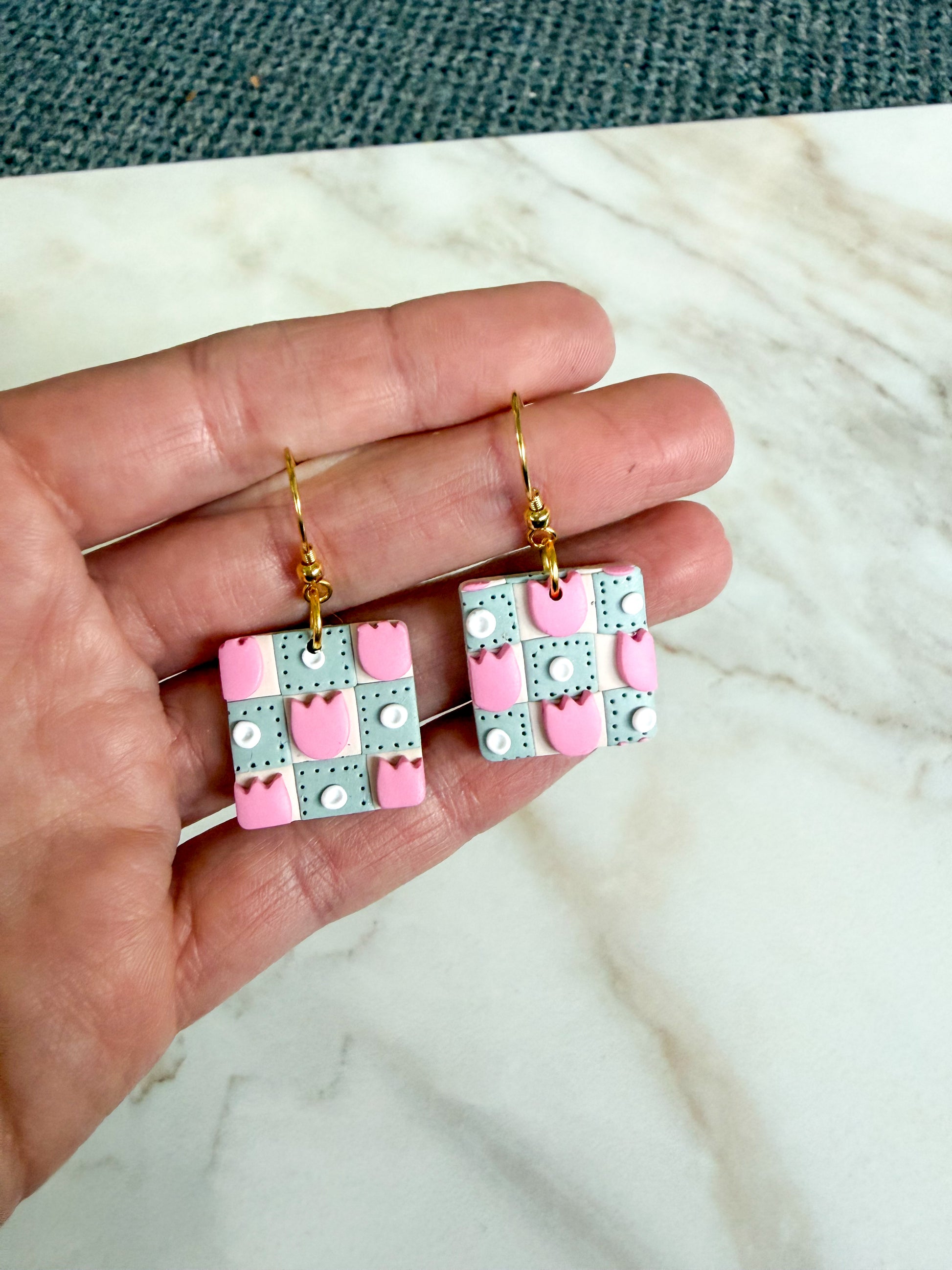 Hand holding square earrings that resemble a green and cream quilt with pink tulips.